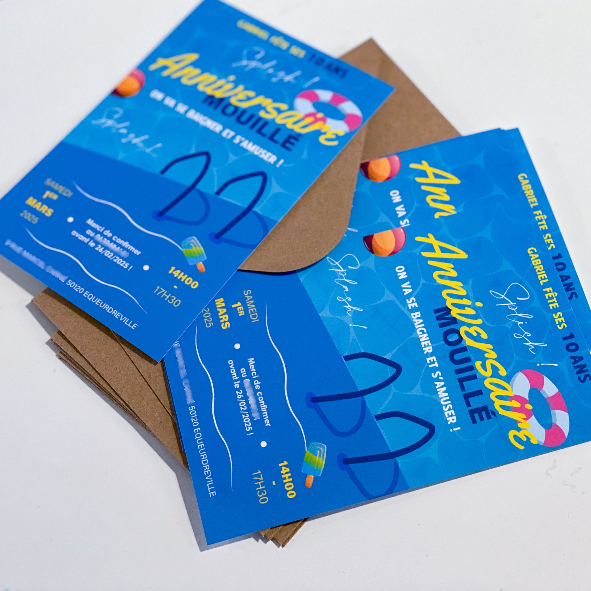 Invitations d’anniversaire thème piscine en bleu vif : motif d’eau en fond, grand titre “Anniversaire” jaune, bouée rayée et ballons, mention “Splash!”, glace, encarts date/heure en bas. Les cartes sont posées sur des enveloppes kraft.