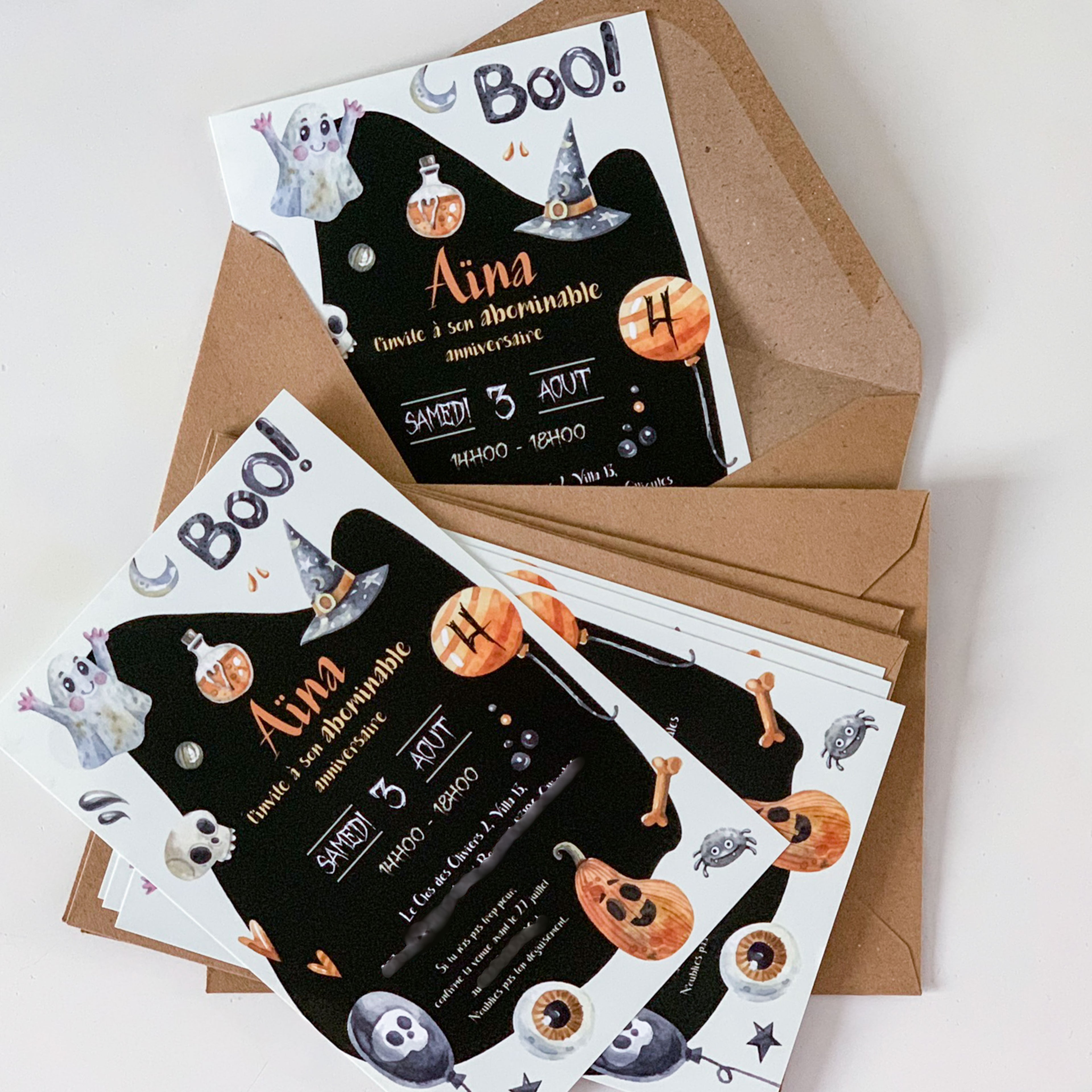 Invitations d’anniversaire thème Halloween posées sur des enveloppes kraft. Fond noir en forme de fantôme, titre “Boo !”, illustrations aquarelle : chapeau de sorcière, fioles, lune, citrouilles, os, araignées et yeux. Prénom “Aina” en orange, date “Samedi 3 août”, horaire “14h00–18h00”.