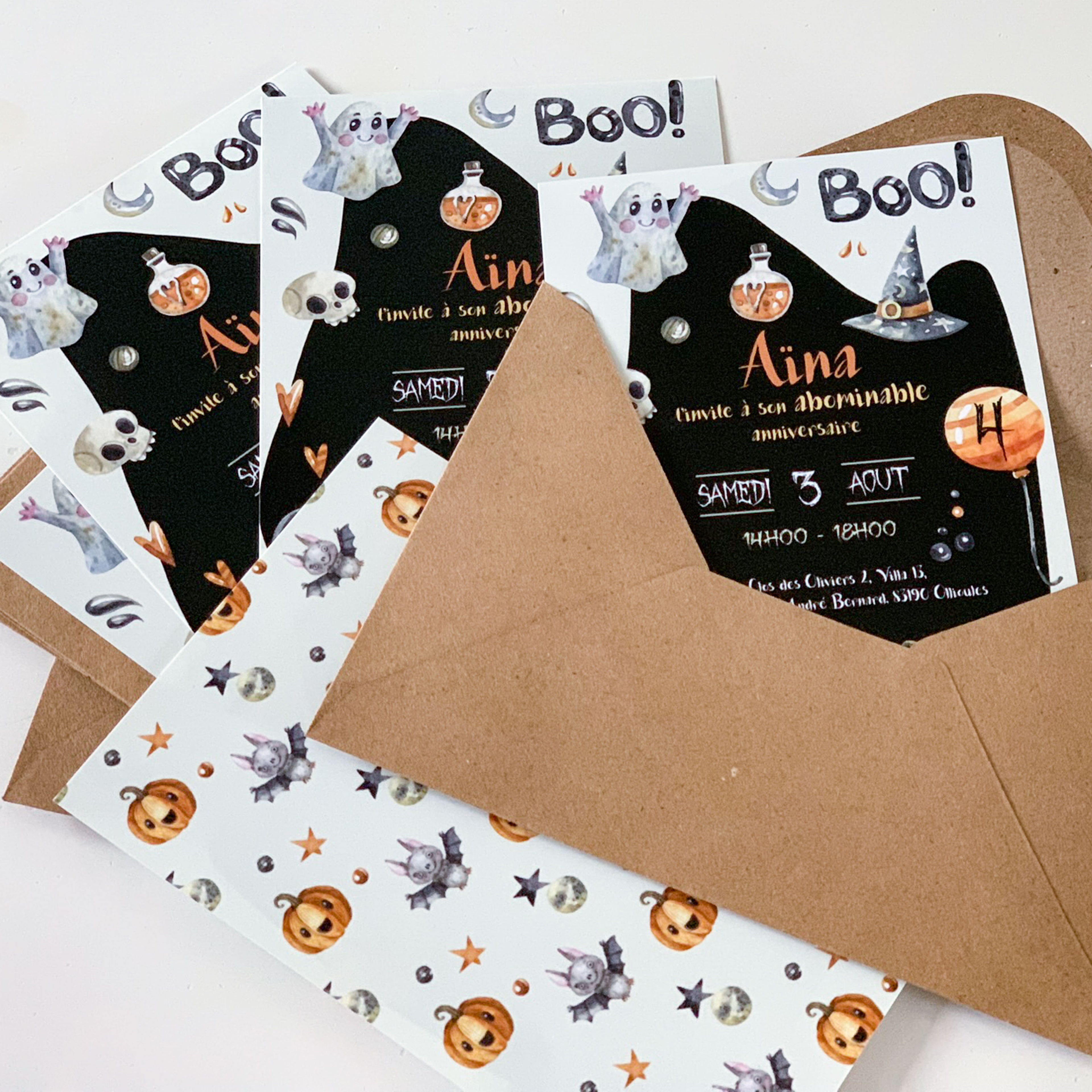 Cinq invitations d’anniversaire imprimées Halloween dont une glissée dans des enveloppes kraft. Fond noir en silhouette de fantôme avec le titre “Boo !”, illustrations aquarelle (chapeau de sorcière, fiole, crâne). Prénom “Aina” en écriture dorée, date “Samedi 3 août”, horaire “14h00–18h00”, ballon orange marqué “4”. En bas, une invitation verso montre un motif citrouilles, lunes, étoiles et petites créatures.