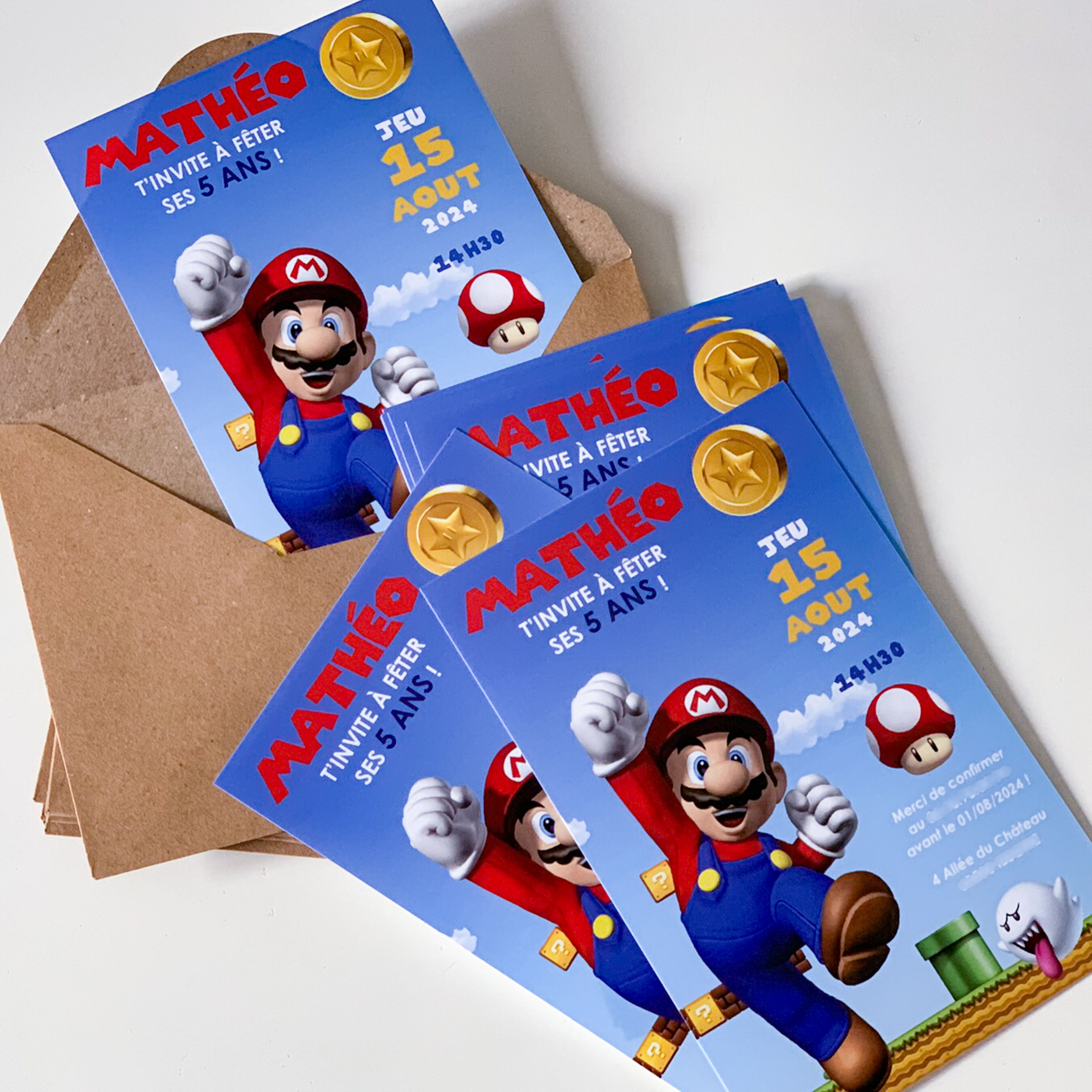 Invitations imprimées Super Mario (Nintendo) : Mario bondit sur fond ciel bleu avec pièces dorées, blocs « ? » et champignon rouge-blanc ; texte “MATHÉO – jeu 15 août 2024 – 14h30”, cartes empilées sur des  enveloppes krafts.
