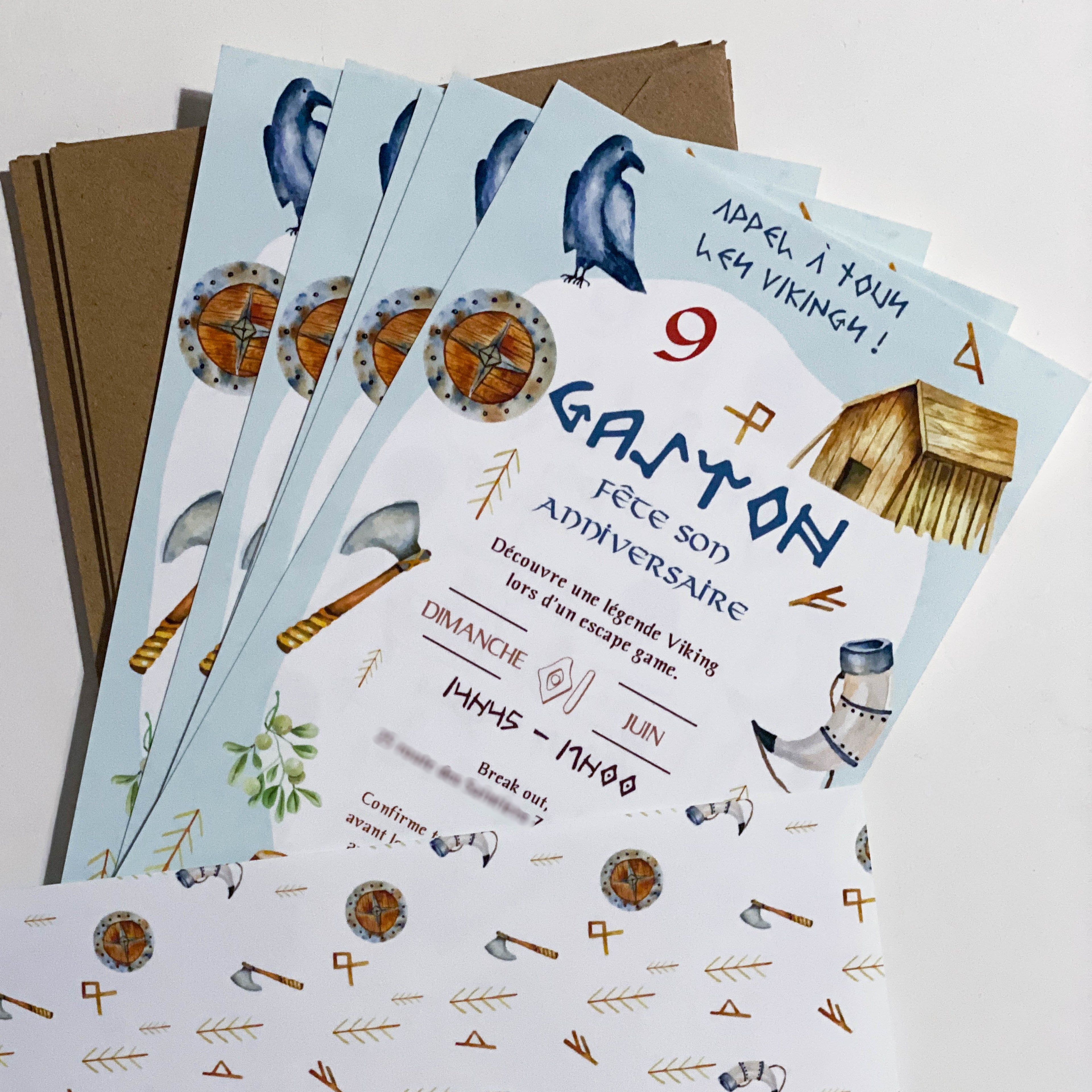 Invitations d’anniversaire thème viking posées en éventail sur des enveloppes kraft. Fond bleu clair avec illustrations aquarellées : corbeau, boucliers ronds bois/métal, haches, corne à boire, flèches et petite maison en bois. Au centre, le prénom “GASTON” en lettres runiques, le chiffre “9”, texte “Appel à tous les Vikings !”, mention d’un escape game, encarts date “dimanche … juin” et horaire “14h45–17h00”. En bas, une enveloppe blanche à motifs de haches, boucliers et flèches.