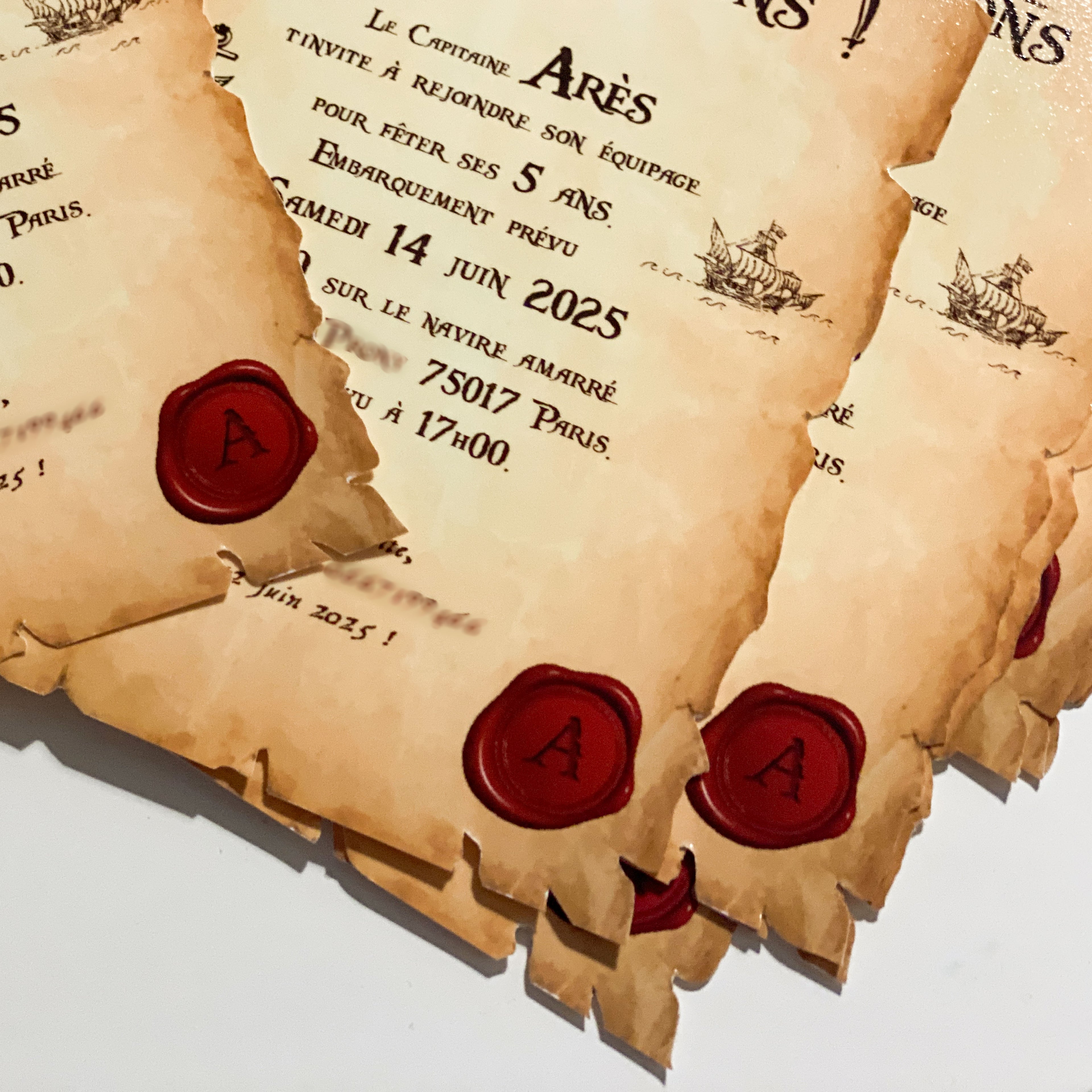 Gros plan sur des invitations pirates effet parchemin vieilli : bords déchirés/brûlés, cachets de cire rouges “A”, petites illustrations de navires, texte personnalisé (« Capitaine Arès fête ses 5 ans – samedi 14 juin 2025 – 75017 Paris – 17h00 »), cartes superposées.