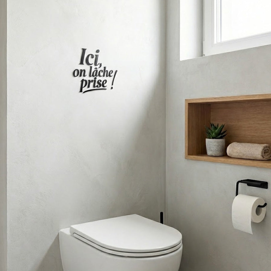 Décoration murale “Ici, on lâche prise !” en impression 3D (PLA), posée sur un mur clair au-dessus de toilettes modernes, avec niche en bois, petite plante et dérouleur papier dans un espace WC lumineux.