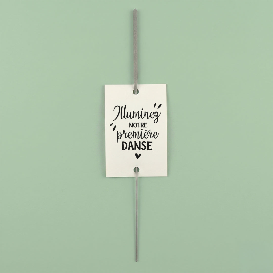Une étiquette rectangulaire blanche "Illuminez notre première danse" avec un petit cœur. Un cierge magique est glissé verticalement au centre de l'étiquette, présentée sur un fond vert clair.