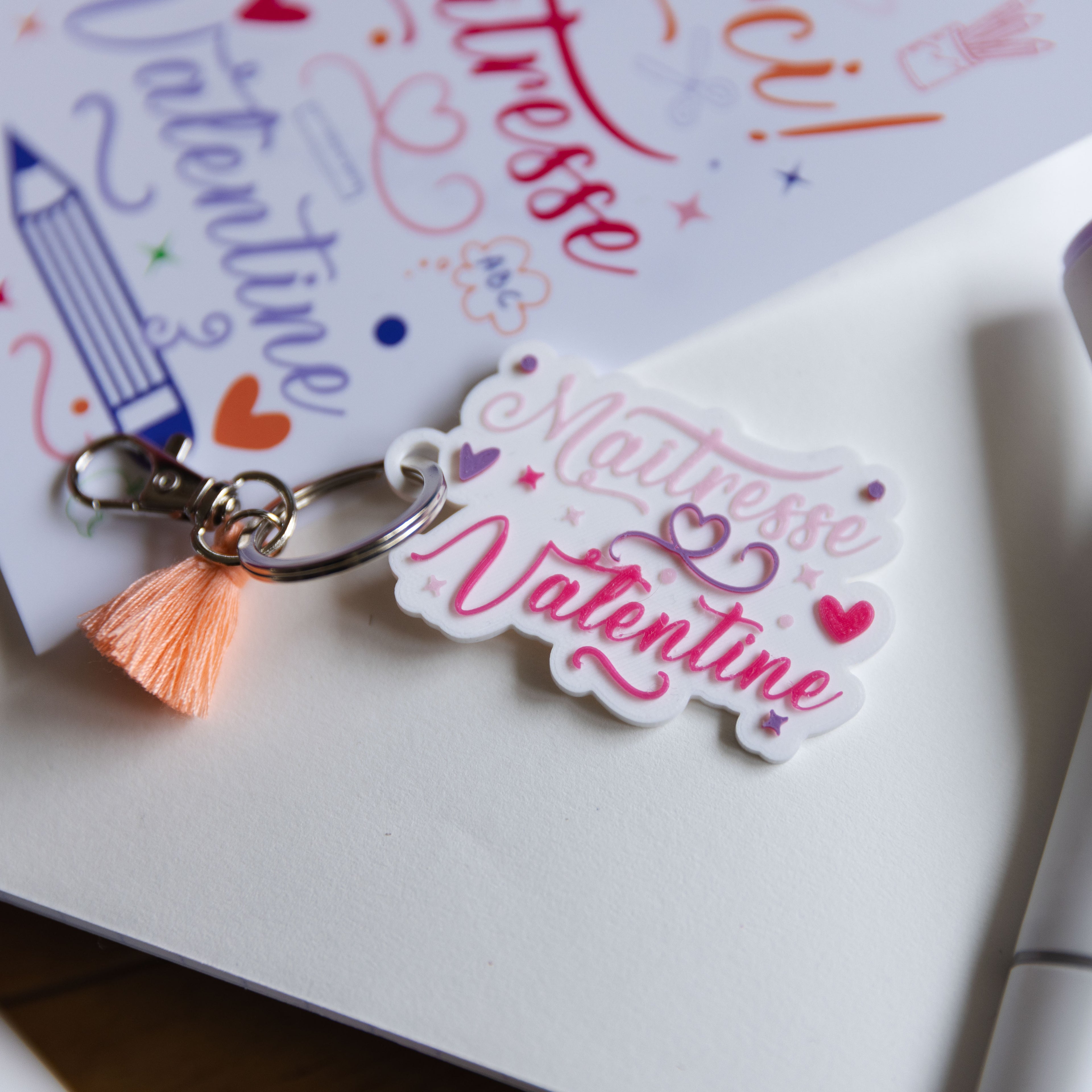 Au centre, en focus parfait, se trouve le porte-clés Maîtresse Valentine identique à l'original, avec son texte en relief, son pompon pêche et son fermoir en métal argenté. 