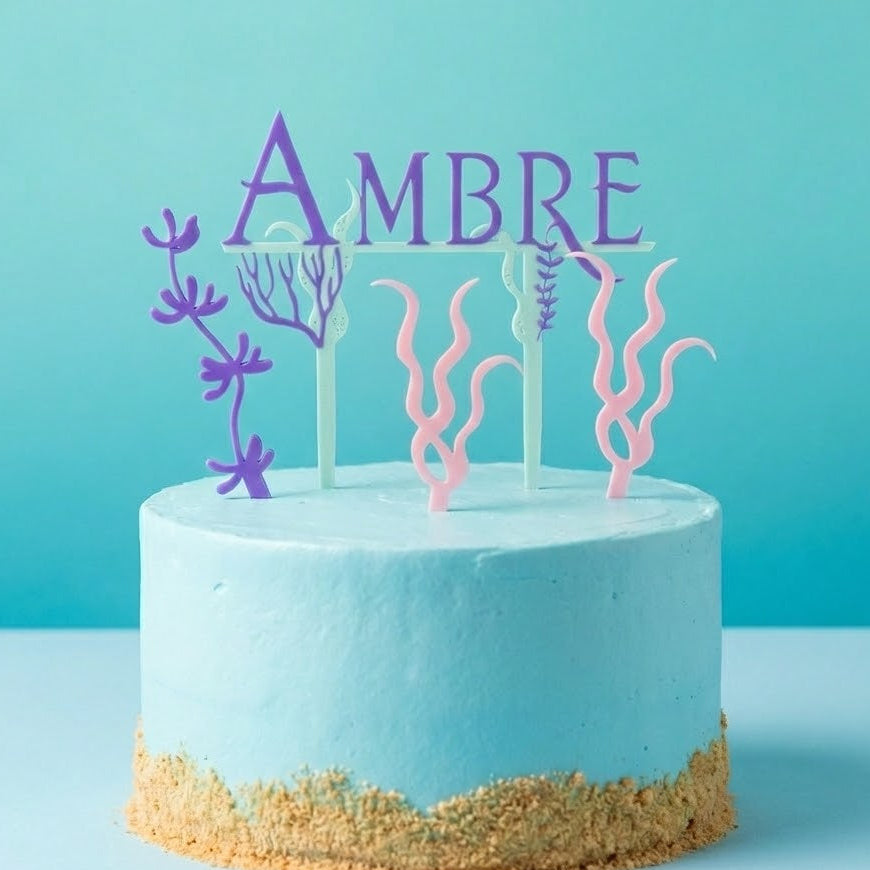 Cake topper prenom Ambre sur un gateau bleu clair, avec decorations marines en forme d algues et coraux roses et violets, theme sirene et ocean, decoration anniversaire enfant.