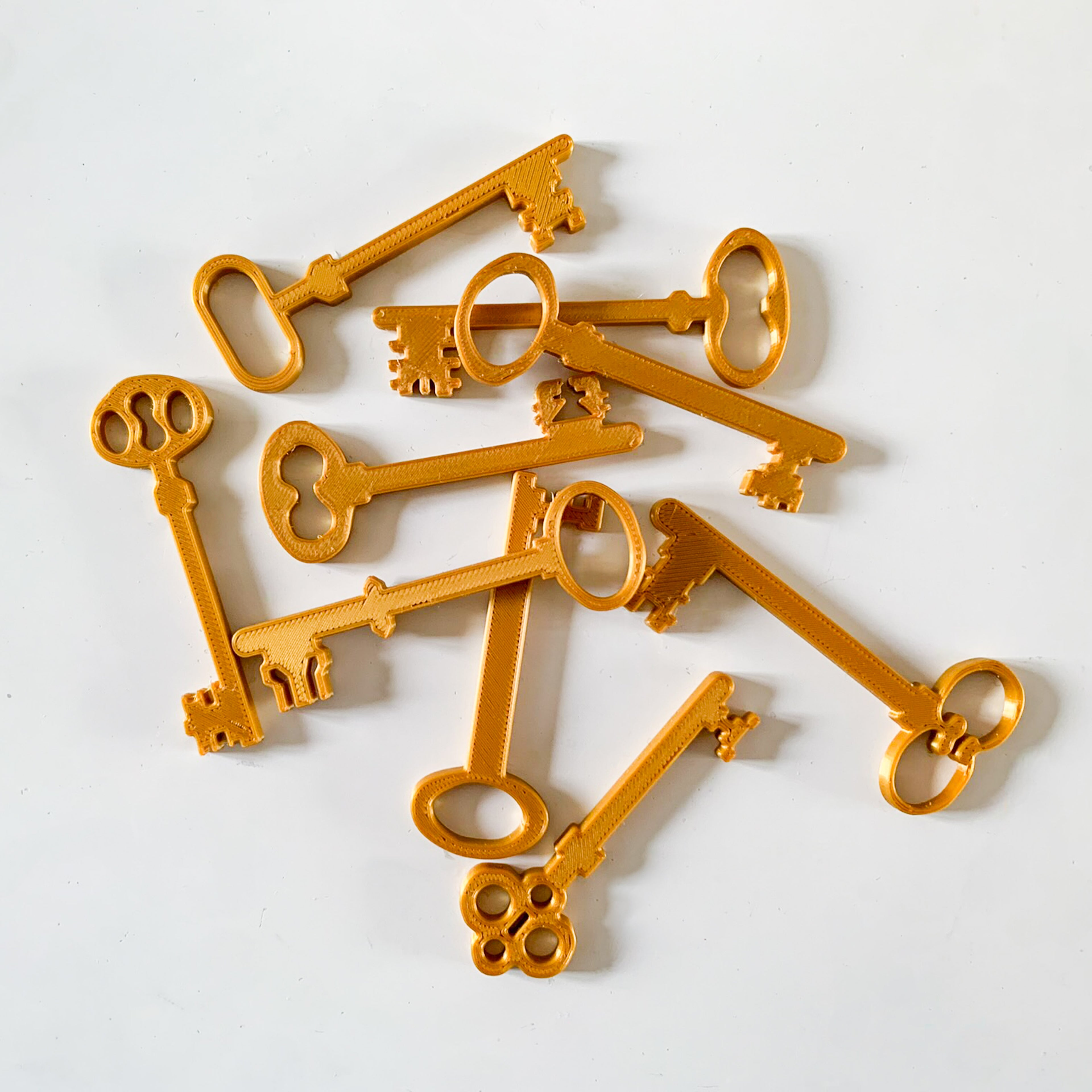 Lot de petites clés anciennes dorées imprimées en 3D en PLA, aux formes vintage variées, disposées en vrac sur un fond blanc, pour servir de décor de table ou d’accessoires de jeu lors d’un anniversaire sur le thème des sorciers, escape game ou chasse au trésor.