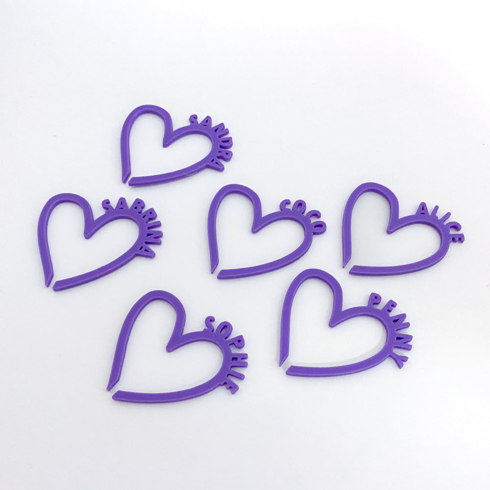 Photo de six marque-verres en forme de cœur, imprimés en PLA violet. Chaque cœur est ouvert à la base pour se clipser sur un verre, et porte un prénom inscrit en lettres fines le long de la courbure droite du cœur. Les pièces sont posées à plat sur un fond blanc, alignées de manière légère et esthétique.