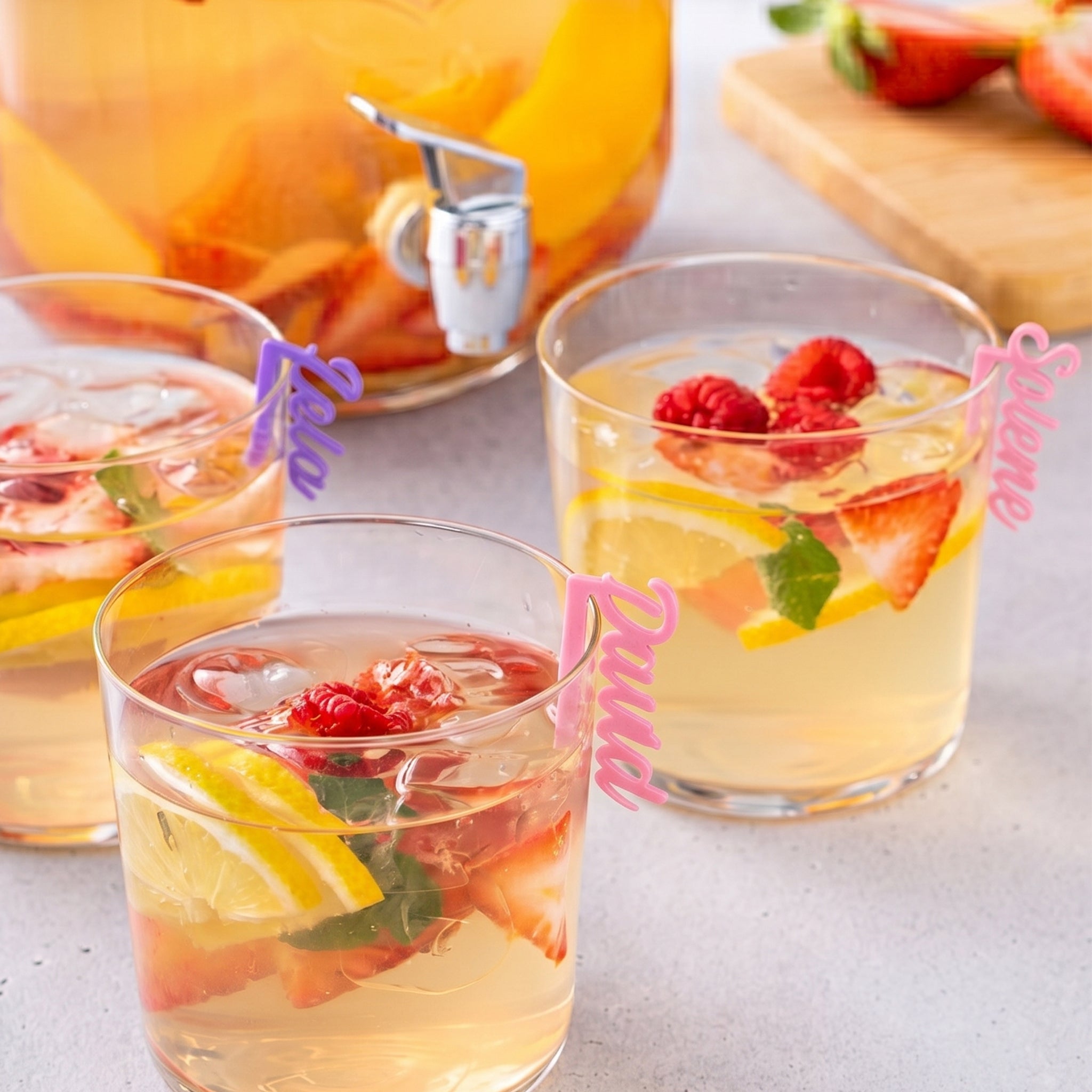 Photographie mettant en vedette un grand distributeur de boissons en verre infusé de fruits (pêches, fraises) avec un robinet. Au premier plan, plusieurs verres assortis sont remplis de glace, de fruits frais (framboises, fraises, tranches de citron) et de menthe. Chaque verre est élégamment orné d'un marque-verre personnalisé et distinctif ("Kela" en violet, "Selene" en rose).