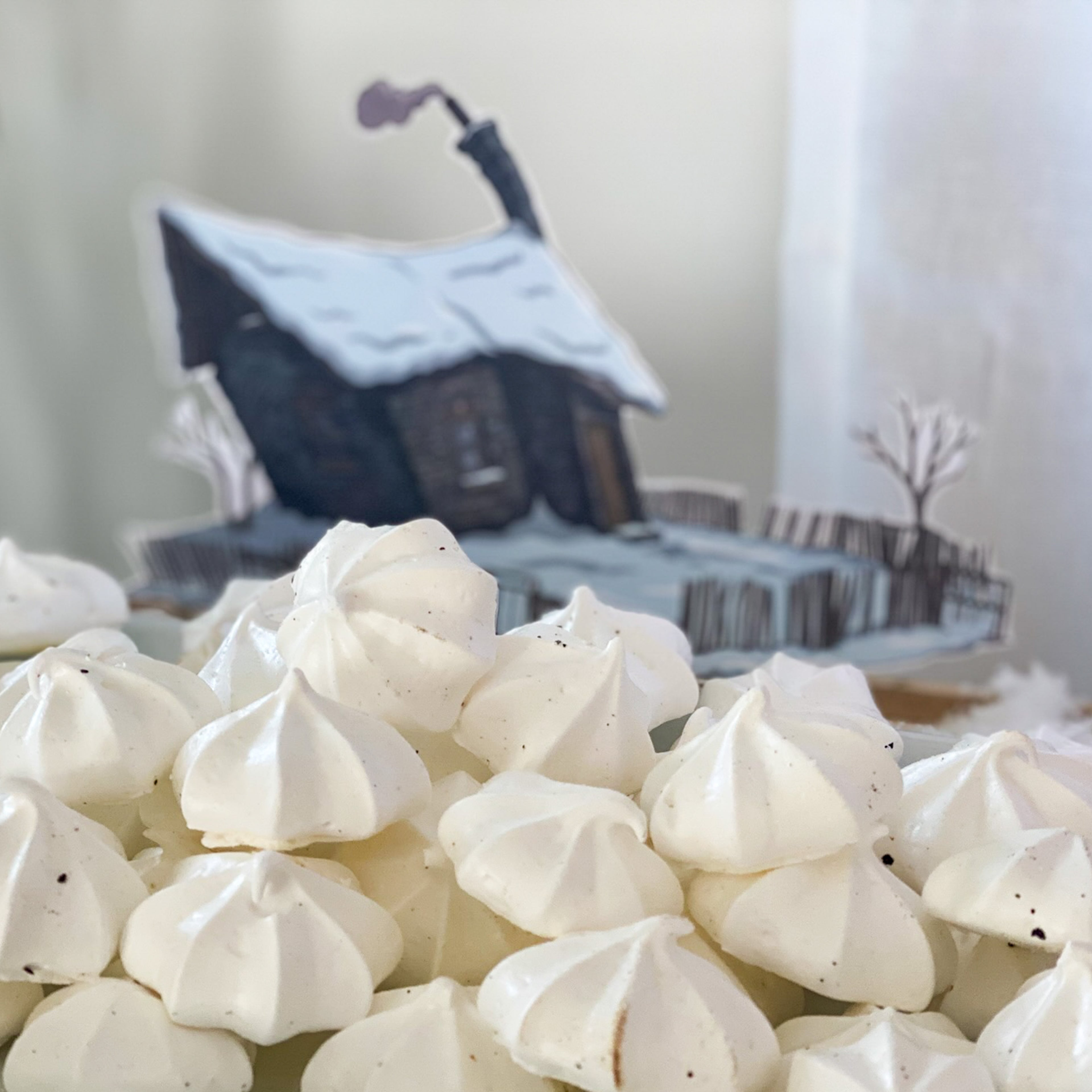 Gros plan sur une montagne de meringues blanches évoquant des sommets enneigés, avec en arrière-plan l'illustration d'une petite maison sous la neige issue du kit PDF. Une mise en scène gourmande qui illustre l'utilisation des décors à imprimer pour une table de fête.