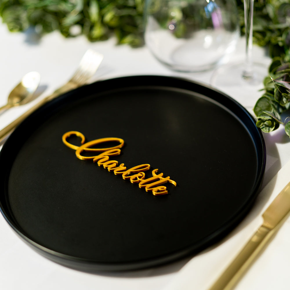 Marque-place calligraphie doré personnalisé « Charlotte », posé sur une assiette noire lors d’une décoration de table élégante.