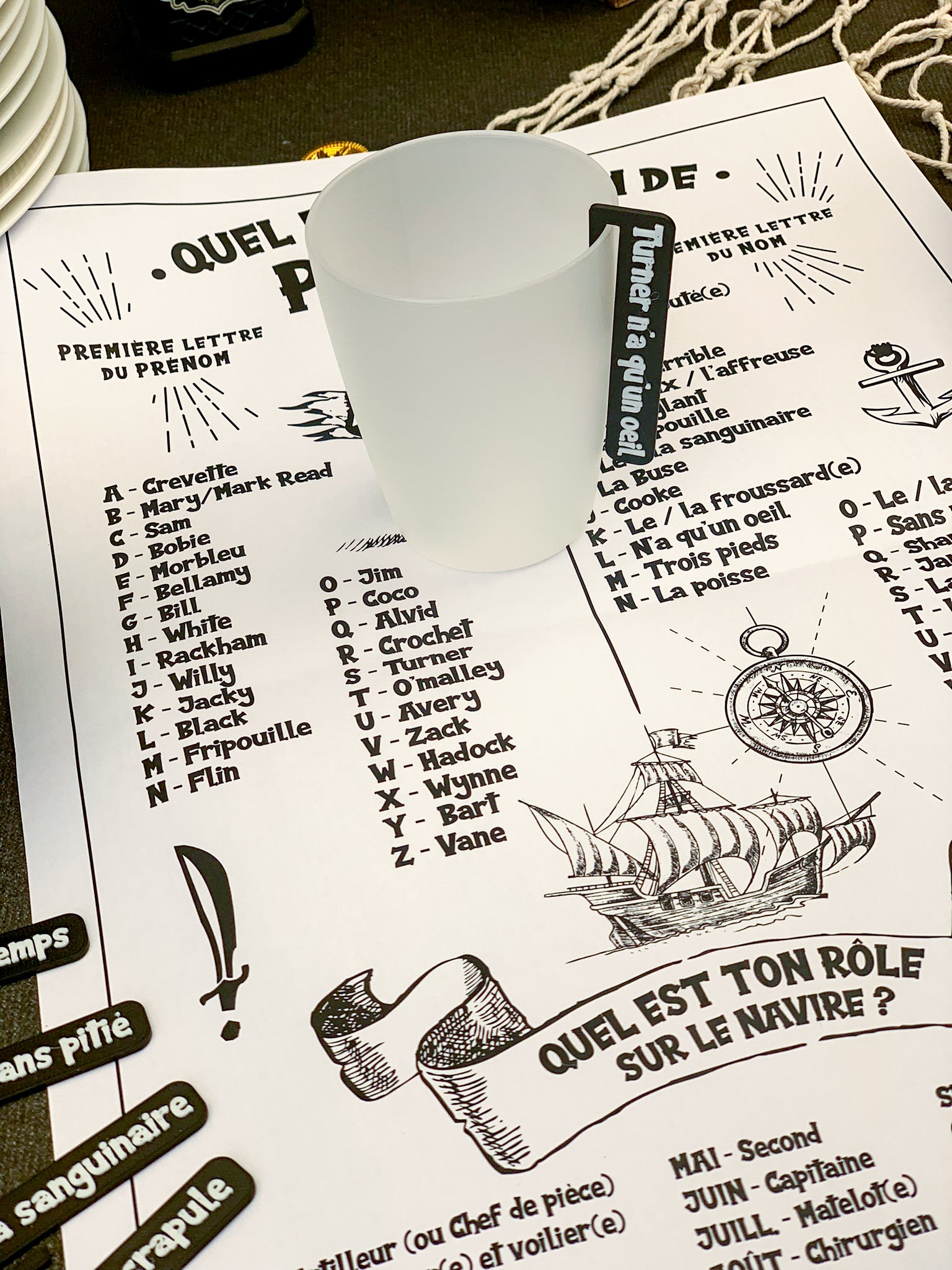Gobelet avec marque-verre Pirate noir posé sur l’affiche “Quel est ton nom de pirate ?”, invitant les invités à chercher leur nom de pirate et à s’immerger dans l’ambiance de la soirée à thème.