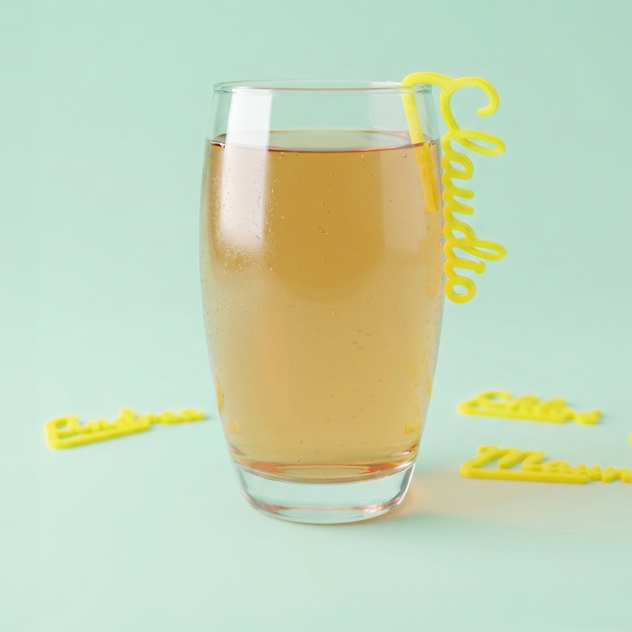 Marque-verre 3D personnalisé jaune avec le prénom Claudio, fixé élégamment sur le rebord d'un verre transparent sur fond vert menthe.