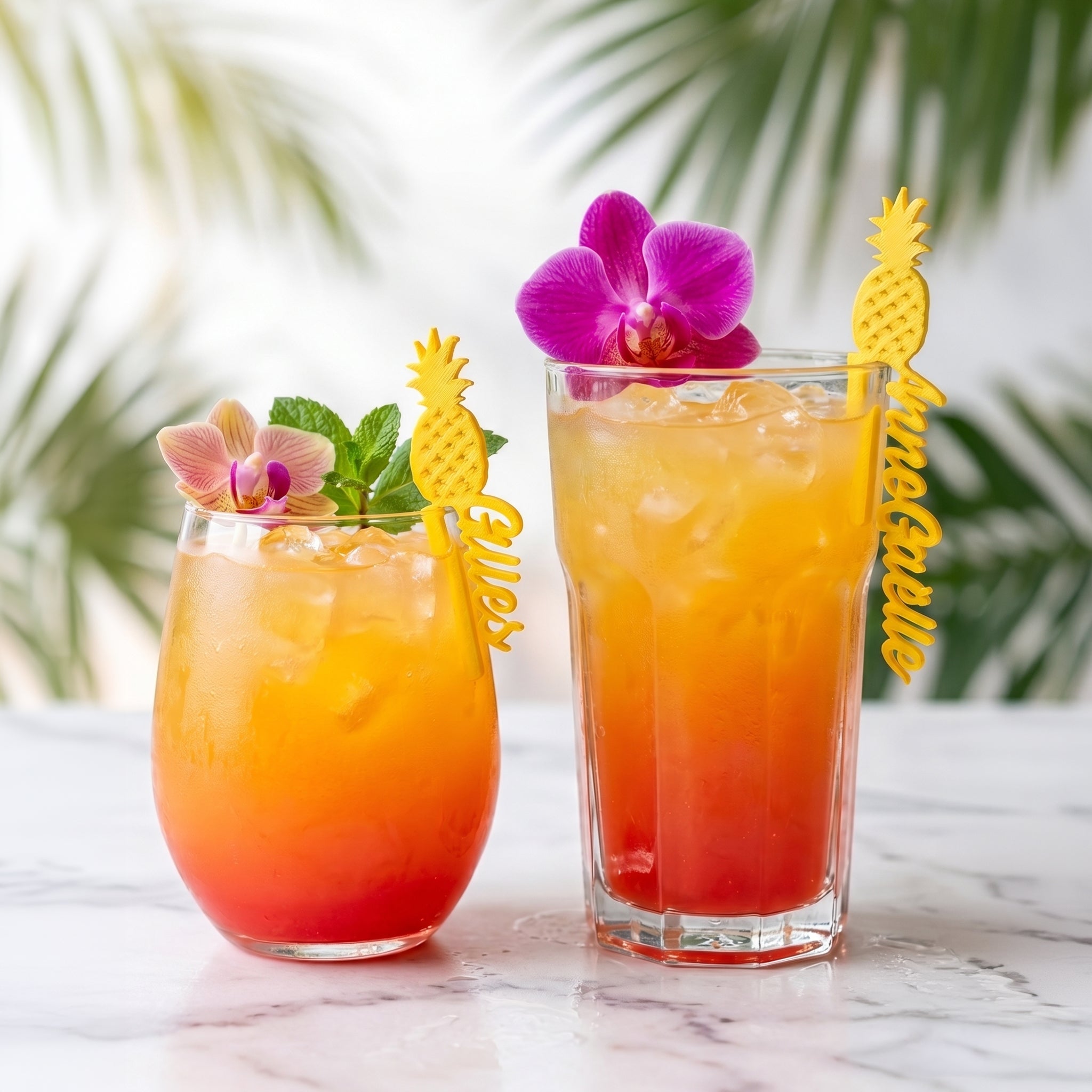 Deux cocktails tropicaux colorés en dégradé d'orange et de rouge (style Tequila Sunrise) reposent sur une table en marbre blanc. Marque-verres personnalisés en forme d'ananas jaune avec les prénoms Kaly et Anne-Gaëlle, clipsés dessus.