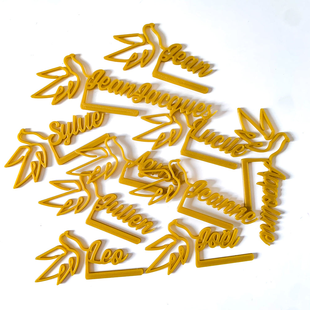 Marque-verres Colombe couleur miel, tous personnalisés avec des prénoms en écriture cursive, disposés en vrac sur fond blanc.