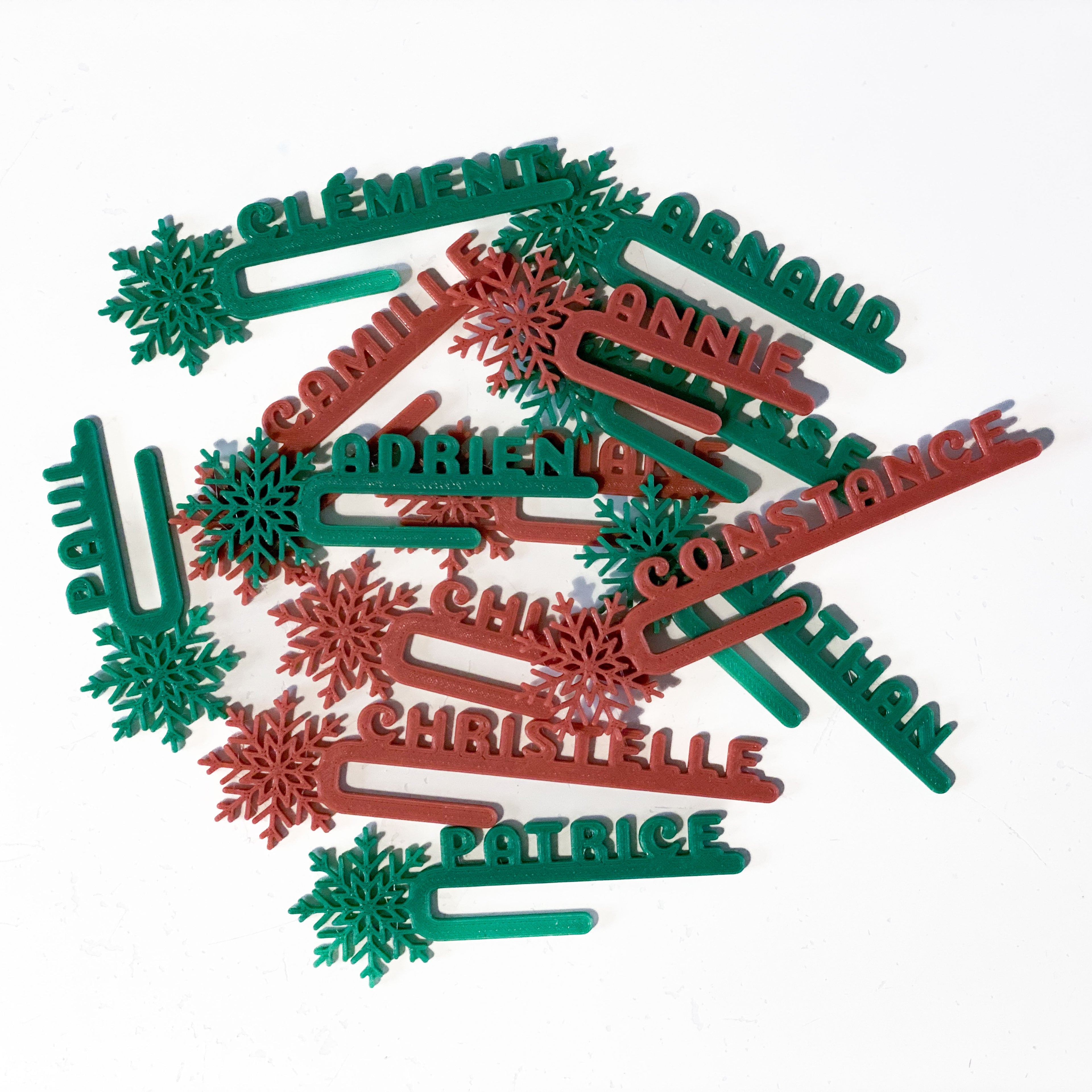 Ensemble de marque-verres Flocon personnalisés en rouge tomette et vert sapin, disposés à plat : chaque pièce présente un prénom et un flocon détaillé pour une décoration hivernale chaleureuse et festive.