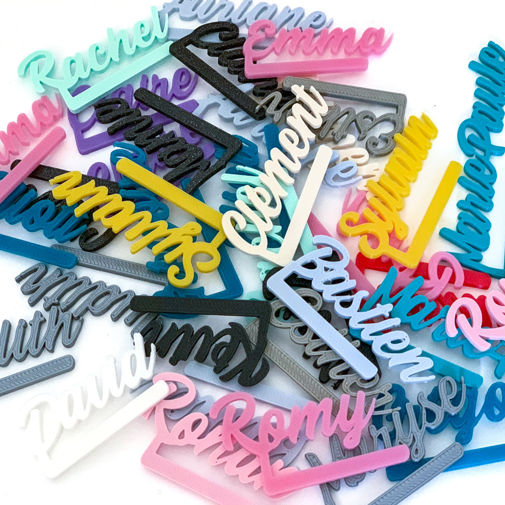 Gros plan sur un ensemble de marque-verres personnalisés en forme de prénoms cursifs, imprimés en PLA et proposés dans de nombreuses couleurs : rose, turquoise, jaune, noir, gris, blanc, bleu et violet. Les pièces sont entassées les unes sur les autres, laissant apparaître plusieurs prénoms comme “Rachel”, “Emma”, “Clément”, “Romy”, “Bastien”, “Sylvain” ou “Paula”. La photo met en valeur la variété des couleurs disponibles et le style cursif caractéristique de la gamme Originels.