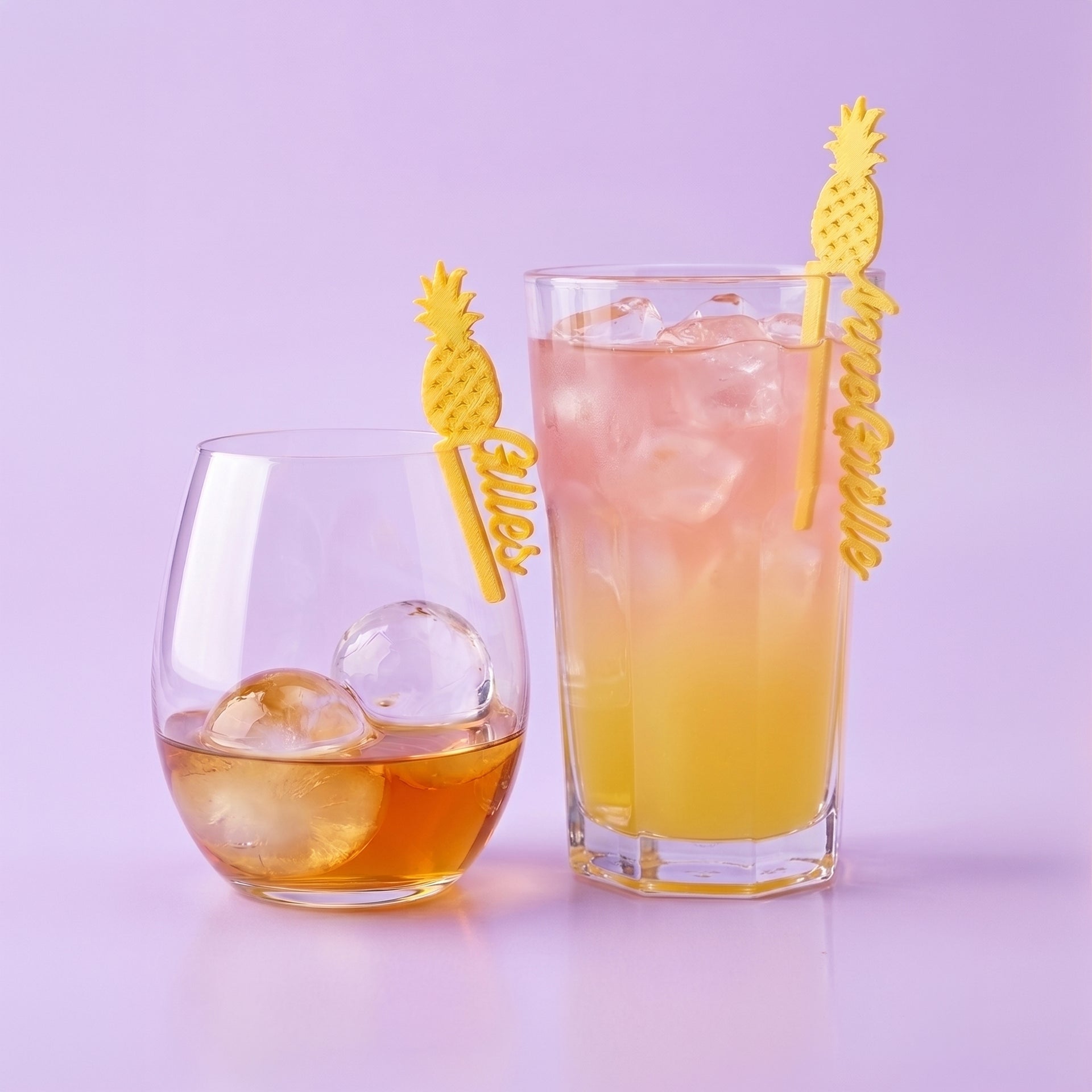 Marque-verres personnalisés en forme d'ananas dorés avec les prénoms Gilles et Anne-Gaëlle, clipsés sur deux verres de cocktail sur un fond lilas uni.