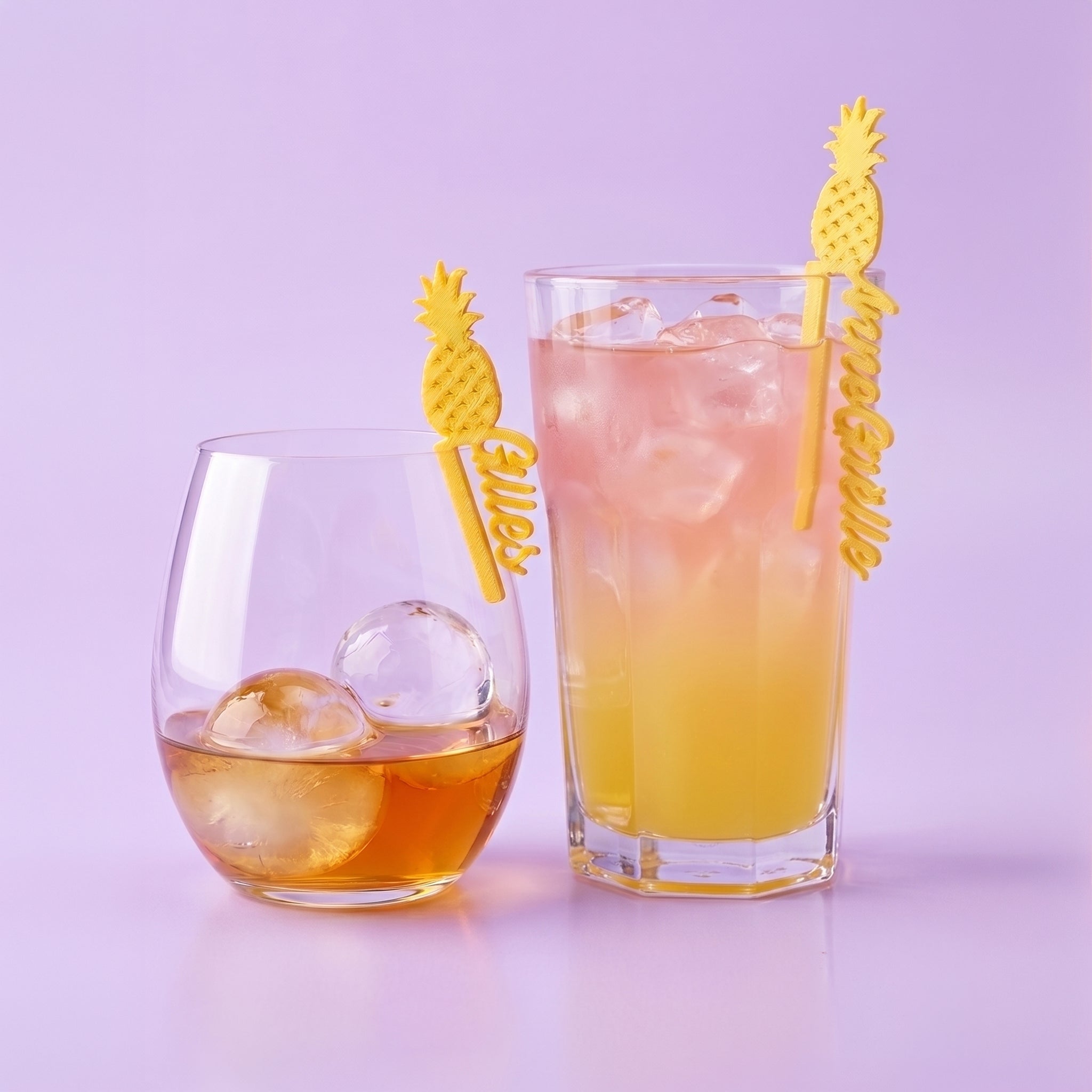 Marque-verres personnalisés en forme d'ananas dorés avec les prénoms Gilles et Anne-Gaëlle, clipsés sur deux verres de cocktail sur un fond lilas uni.