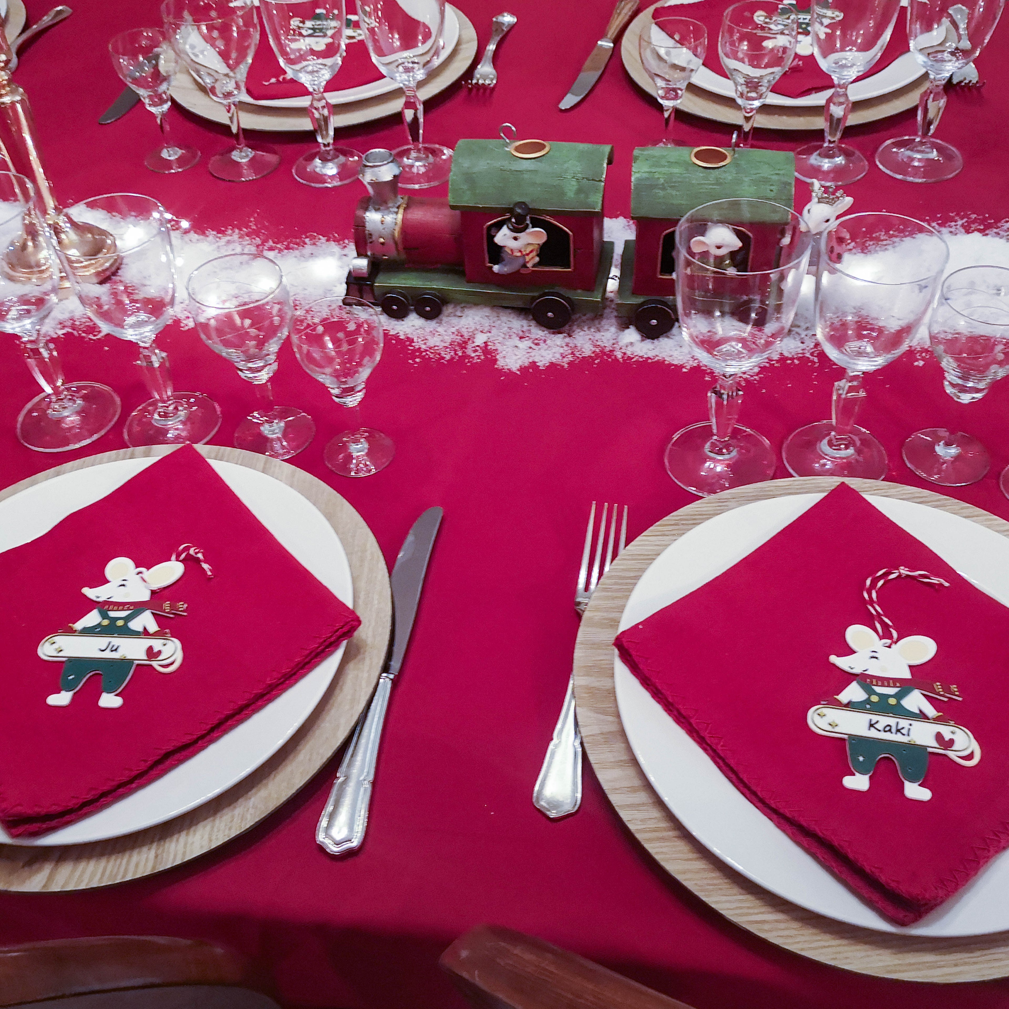 Table de fête de Noël dressée : deux assiettes avec serviettes rouges et marque-places souris personnalisables avec prénoms (ex. “Ju”, “Kaki”), nombreux verres et couverts, avec un petit train décoratif et effet neige au centre de table.