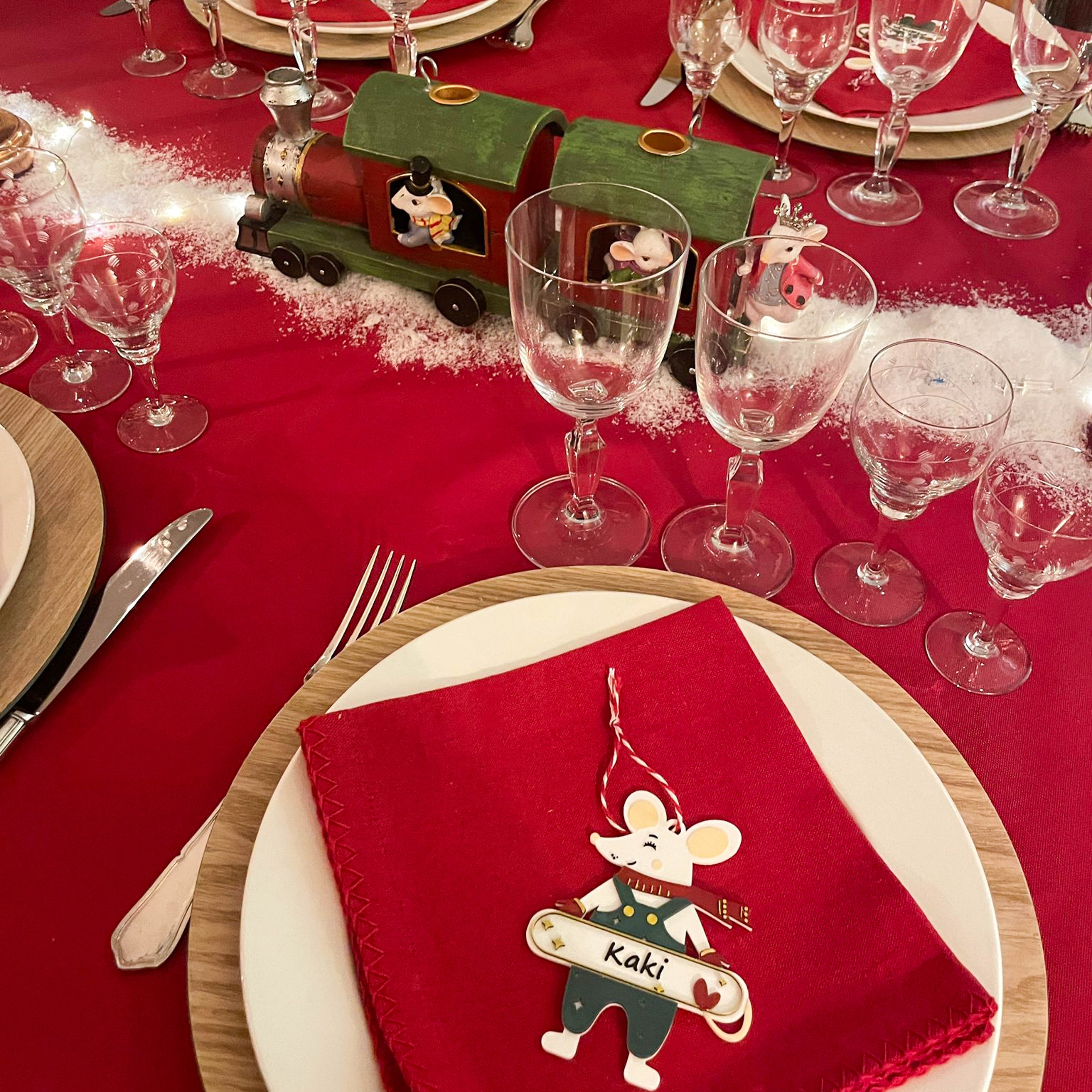 Décoration de table de Noël : marque-place en forme de souris personnalisable avec prénom (ex. “Kaki”), posé sur une serviette rouge dans une assiette, avec une table de fête dressée (verres, couverts) et un petit train décoratif en arrière-plan.