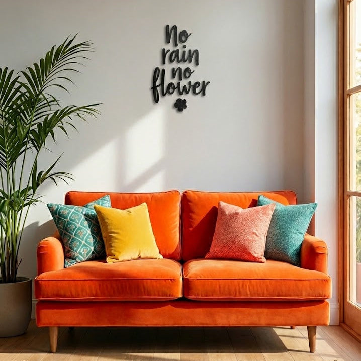 Mise en scène d’une décoration murale “No rain no flower” en impression 3D (PLA) noire, fixée sur un mur clair au-dessus d’un canapé orange avec coussins colorés, près d’une grande plante et d’une fenêtre.