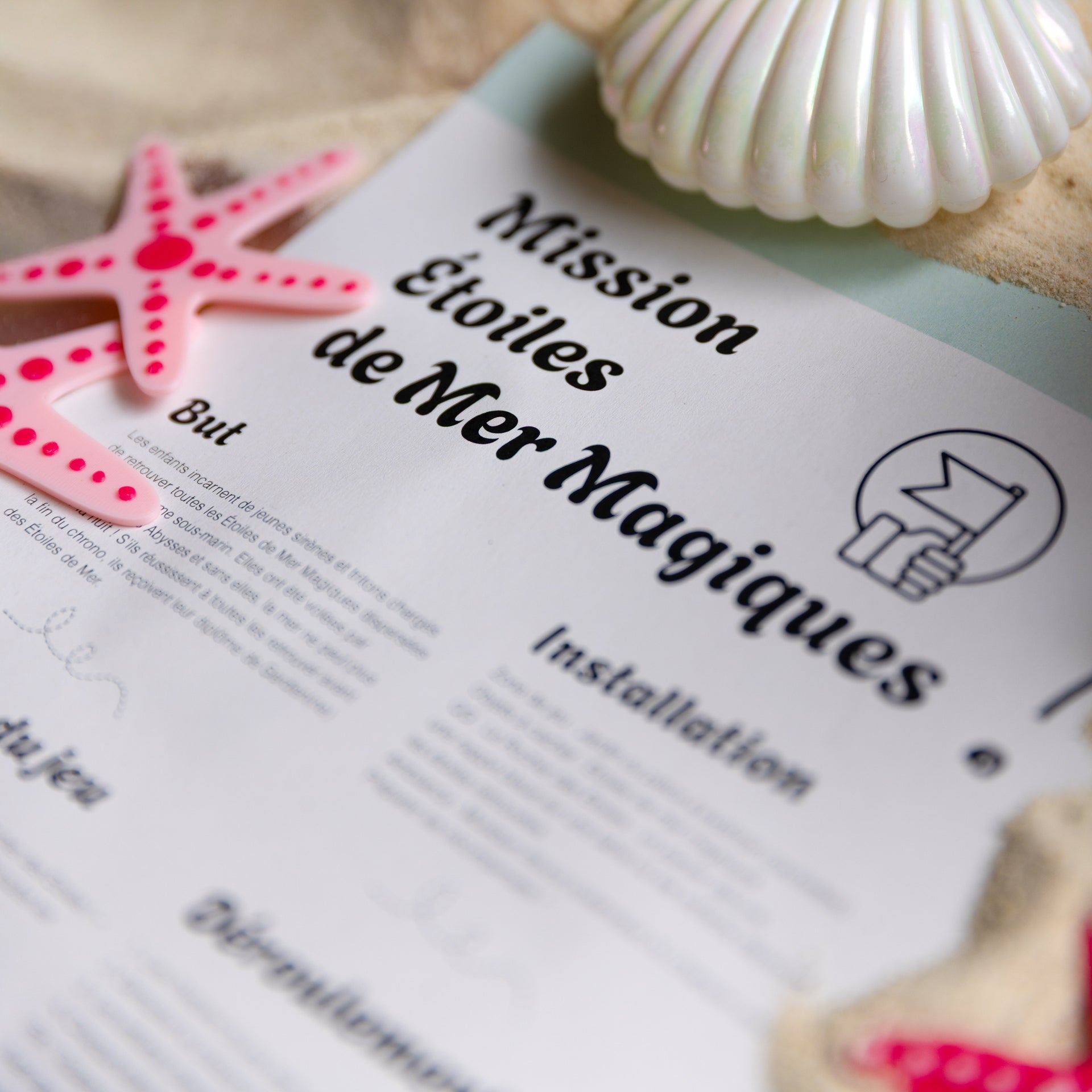 Notice du jeu "Mission Étoiles de Mer Magiques" accompagnée d'étoiles de mer bicolores rose et fuchsia en impression 3D, mises en scène sur du sable pour une chasse au trésor immersive.