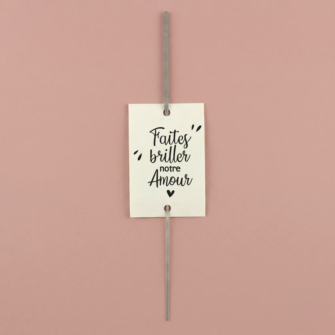 Gros plan d'une étiquette blanche perforée "Faites' briller notre Amour" avec un cierge magique inséré, sur fond vieux rose.