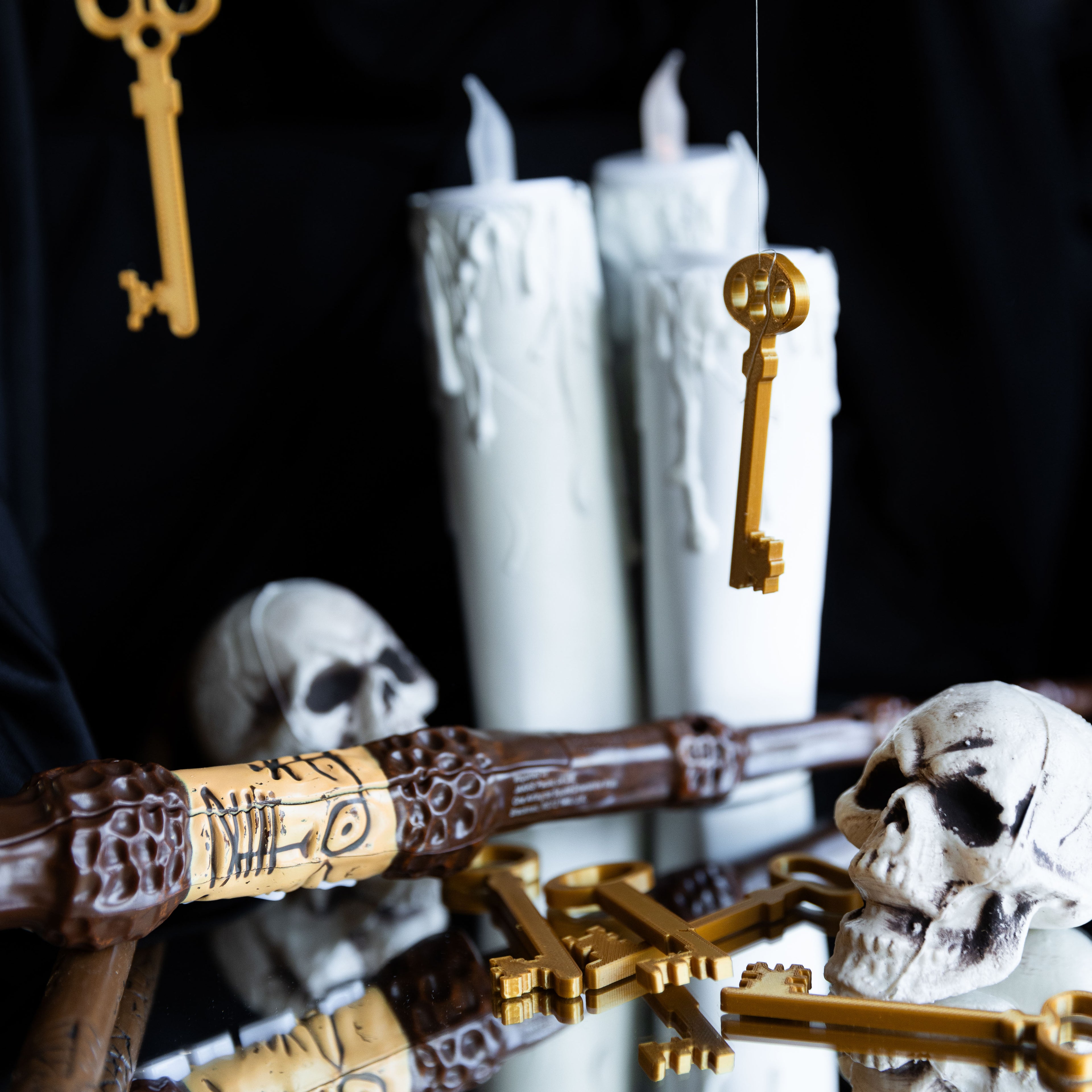 Petit décor de fête sur le thème des sorciers : plusieurs petites clés dorées imprimées en 3D en PLA posées sur un miroir, avec des crânes blancs, une baguette magique sculptée et de grandes bougies blanches fondues sur fond noir, pour illustrer des clés décoratives idéales pour un anniversaire Harry Potter ou Halloween.