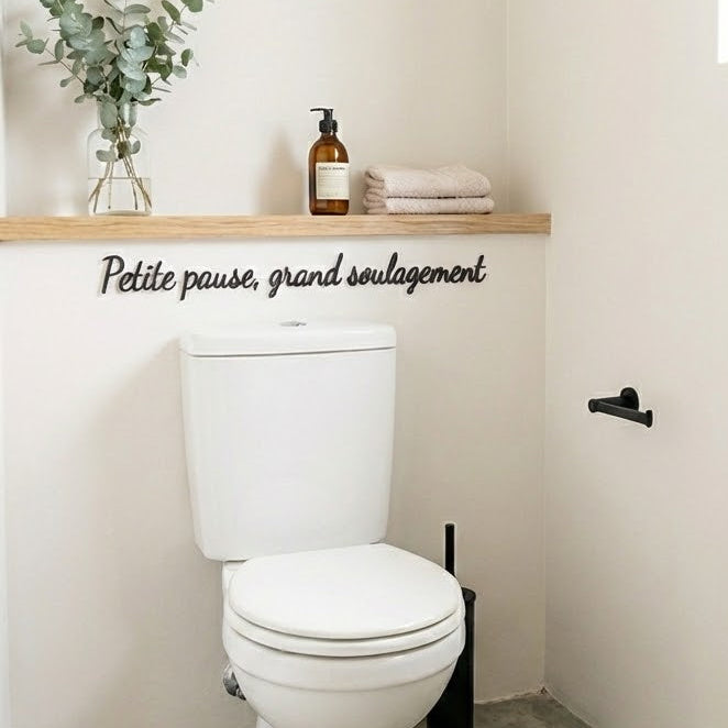 Décoration murale “Petite pause, grand soulagement” en impression 3D (PLA) noire, installée dans des toilettes claires au-dessus du réservoir, avec étagère en bois, serviettes pliées et flacon, style minimaliste.