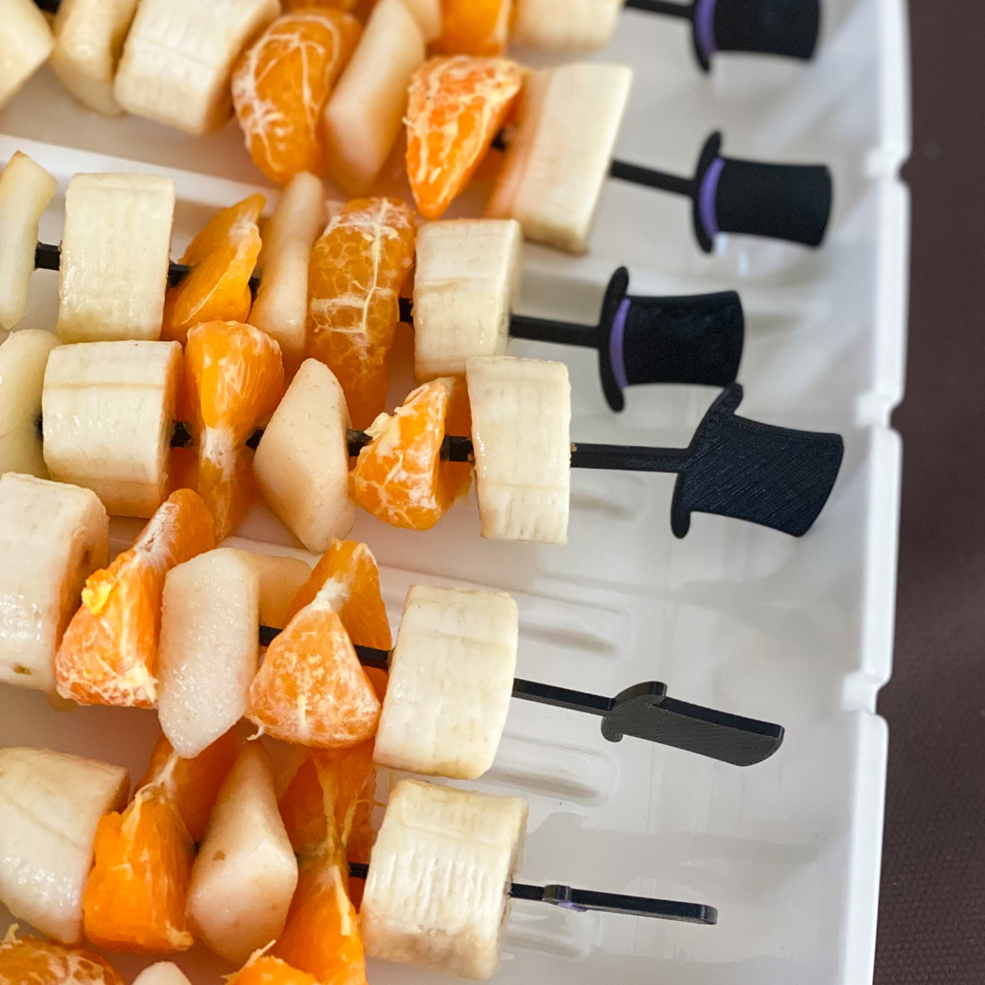 Gros plan sur un plateau de service blanc avec des rangées ordonnées de brochettes de fruits frais. Les piques noires se terminent par de petits chapeaux haut-de-forme noirs liseré décoratifs. Les brochettes alternent rondelles de banane, quartiers de mandarine et dés d'ananas. La texture juteuse des fruits est bien visible. Une présentation élégante pour un buffet de fête.