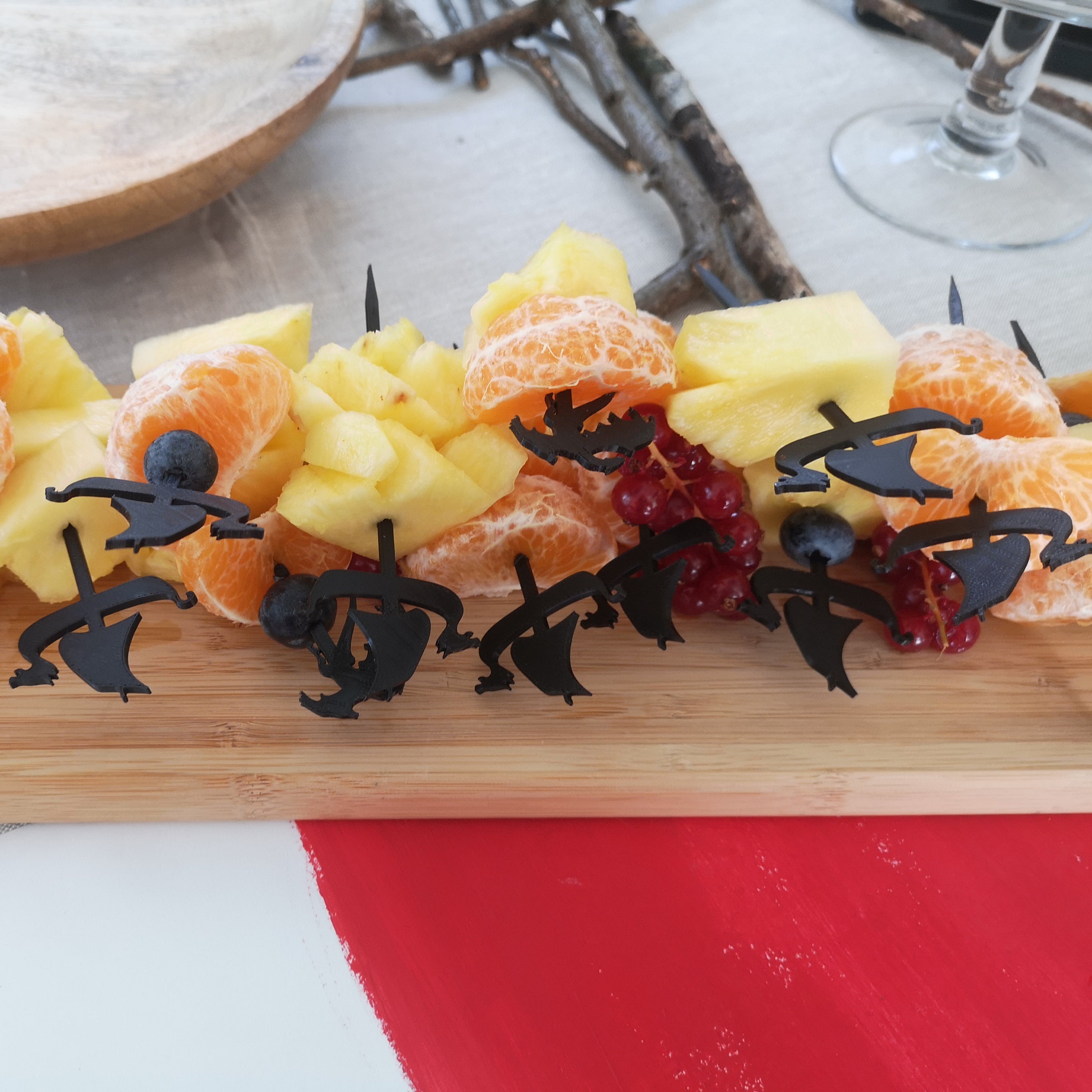 Mise en situation d'un buffet thématique viking présentant des piques à mini-brochettes en forme de drakkar noir. Les piques sont garnies de morceaux de fruits frais, tels que de l'ananas, des clémentines et des myrtilles, et sont disposées sur un plateau en bois.