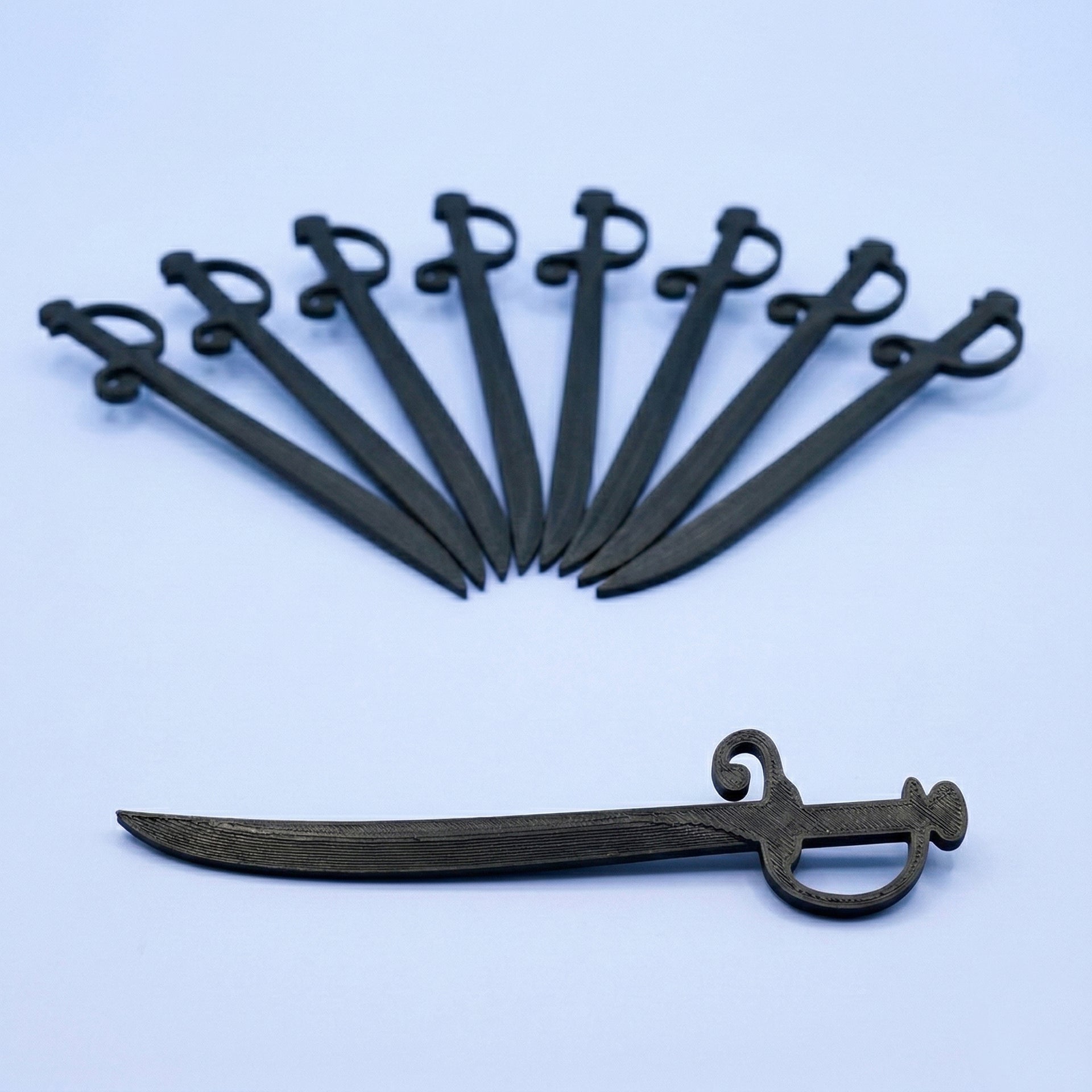 Piques apéritif en forme de sabres de pirate noirs imprimés en 3D disposés en éventail sur un fond bleu. Accessoire de décoration pour buffet d'anniversaire thématique.