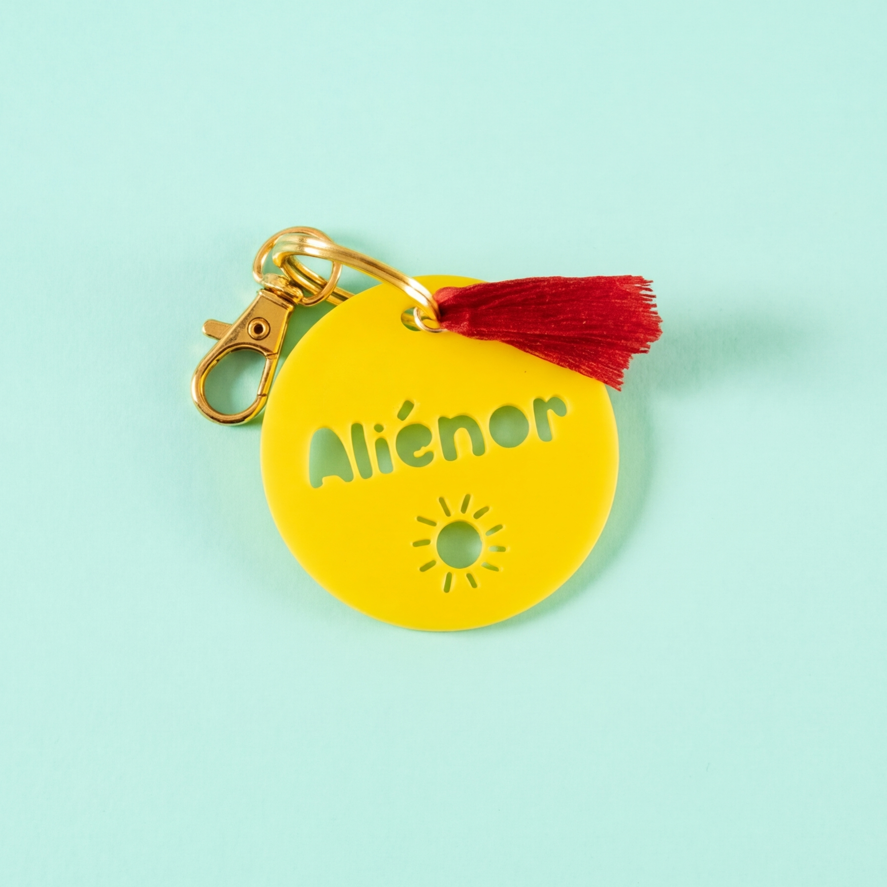 Une vue de dessus d'un porte-clés personnalisé en PLA jaune rond, avec le nom « Aliénor » et un motif de soleil. Il est attaché à un anneau et un mousqueton dorés, avec un petit pompon rouge vif, sur un fond uni menthe à l'eau.