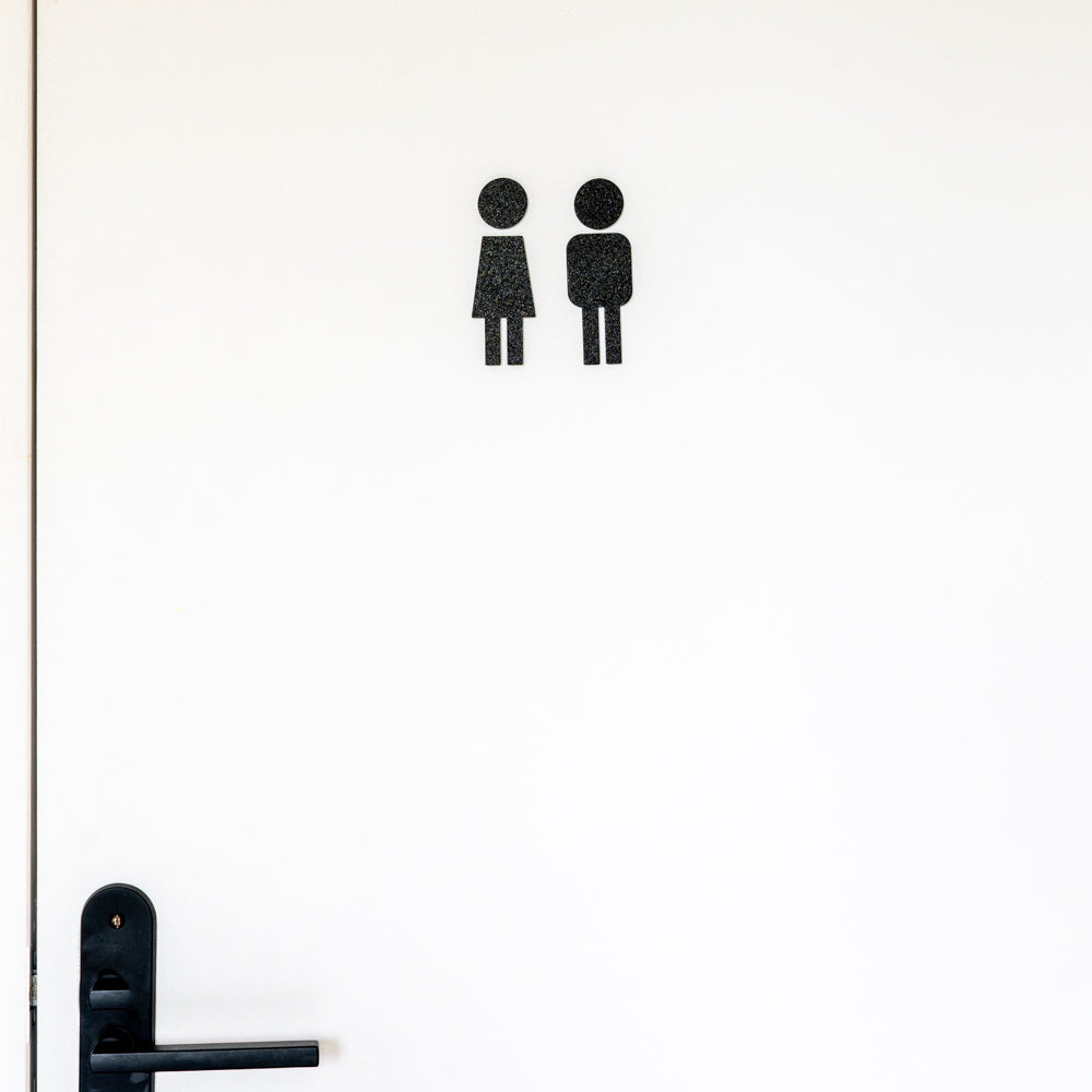 Porte blanche de toilettes avec deux pictogrammes noirs stylisés fille et garçon en relief au centre et poignée noire en bas à gauche, signalétique familiale WC mixte au design minimaliste.