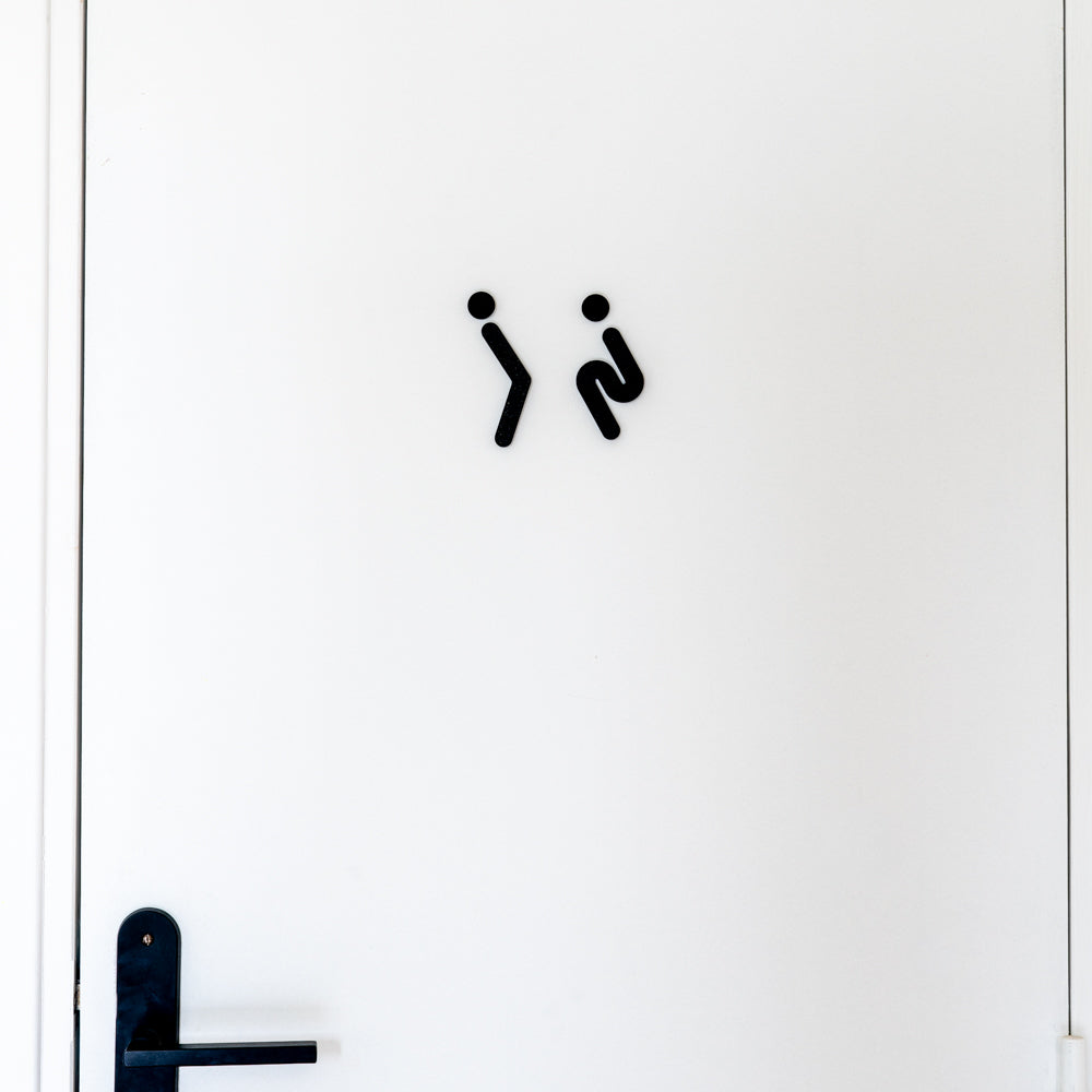 Porte blanche de toilettes avec pictogrammes noirs humoristiques d’un homme et d’une femme qui se retiennent d’uriner, au-dessus d’une poignée noire, signalétique rigolote pour WC.