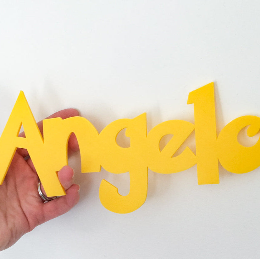 Gros plan sur le prenom Angelo en lettres 3D jaunes en PLA, tenu a la main devant un mur blanc, decoration murale personnalisee pour chambre d enfant.