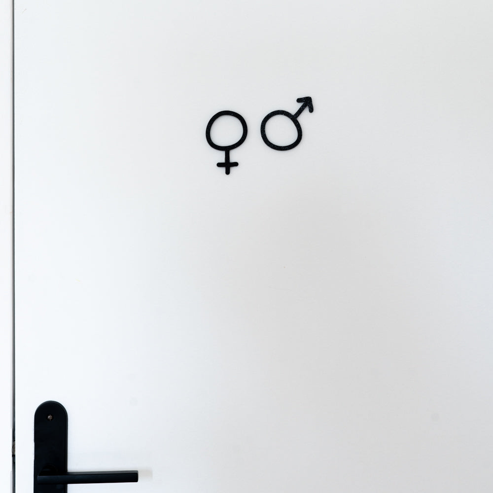 Signalétique toilettes brut fixée sur une porte blanche, avec les pictogrammes noirs homme et femme au centre et une poignée de porte noire en bas à gauche, pour indiquer clairement