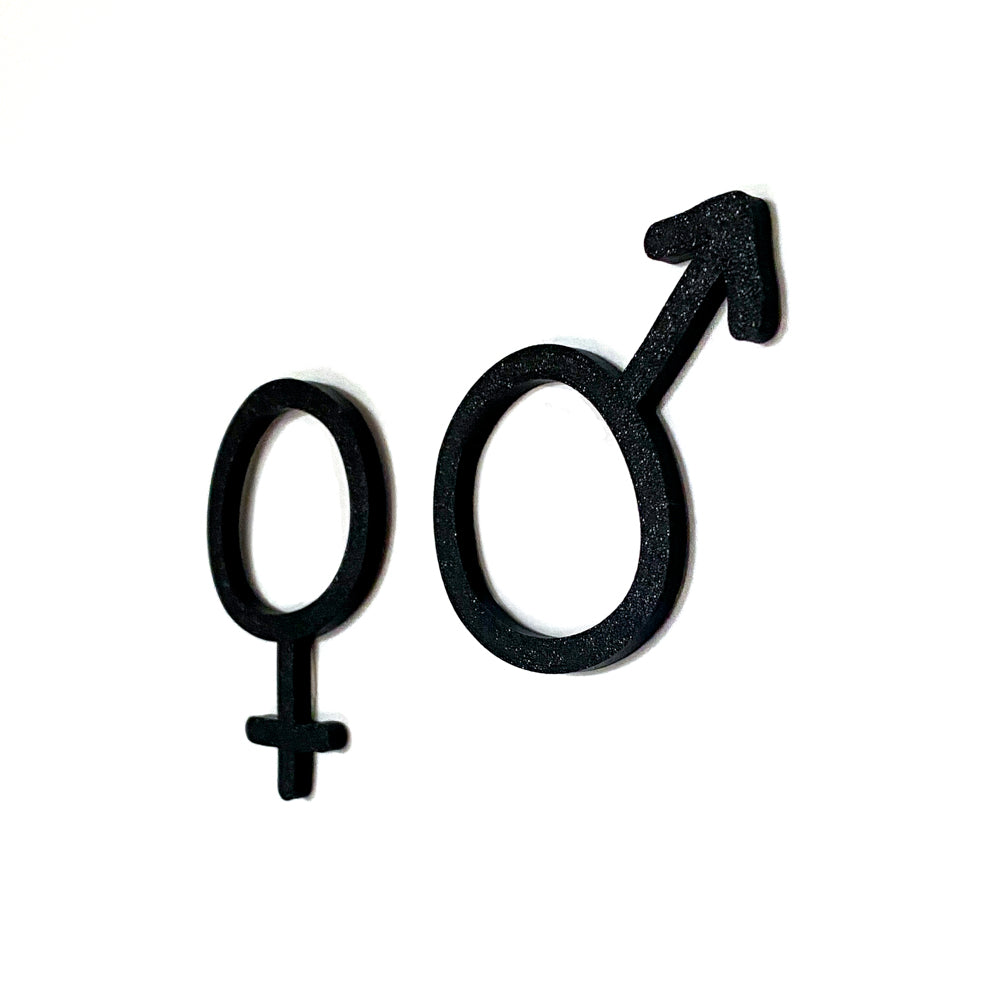 Signalétique toilettes en style brut avec deux pictogrammes noirs, symbole féminin et symbole masculin en relief, posés sur fond blanc, idéale pour indiquer les sanitaires dans une maison, un café ou un lieu d’événement.