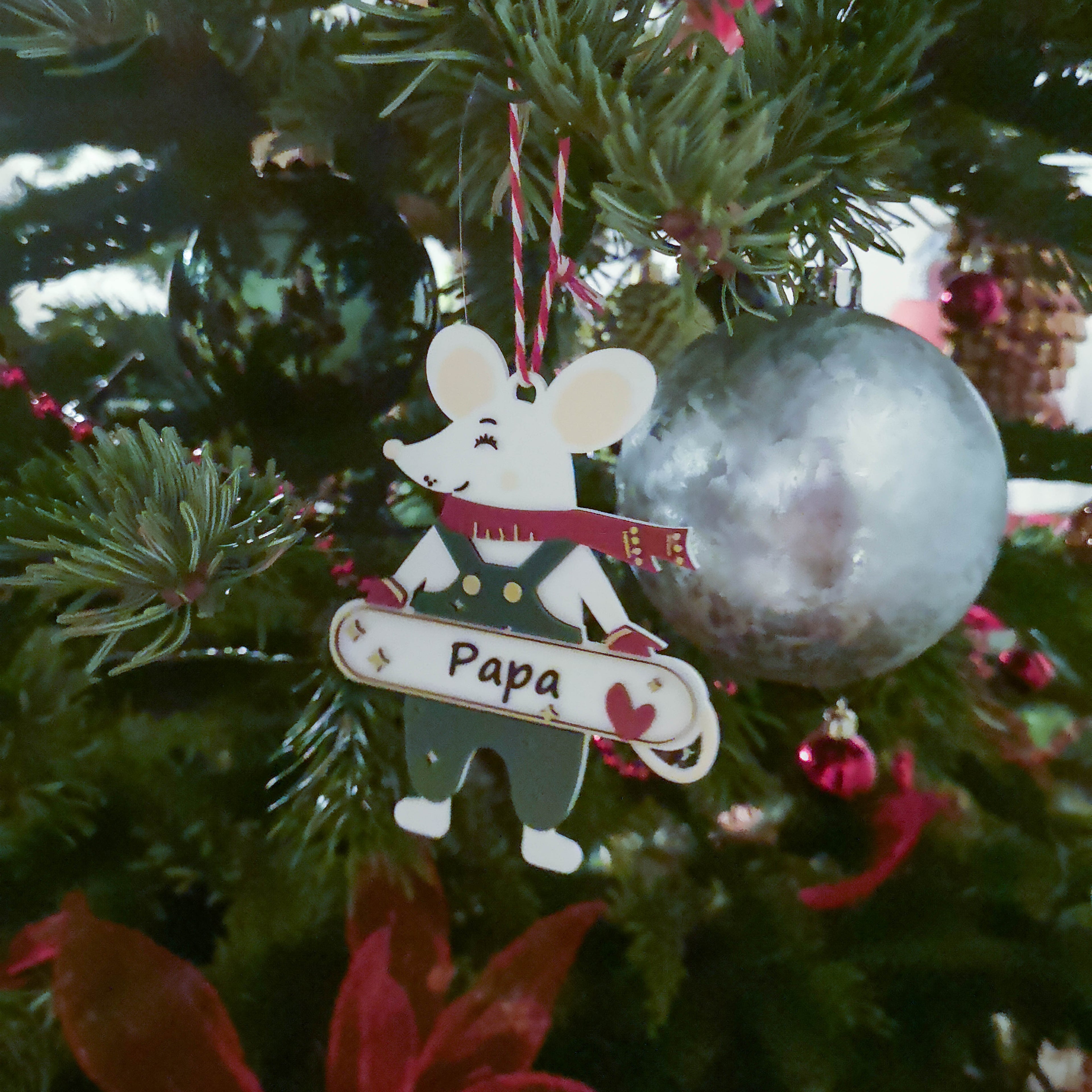Décoration de Noël personnalisable en forme de petite souris, suspendue sur un sapin : souris avec écharpe et salopette, tenant une pancarte “Papa” avec un cœur, au milieu de boules et guirlandes de Noël.