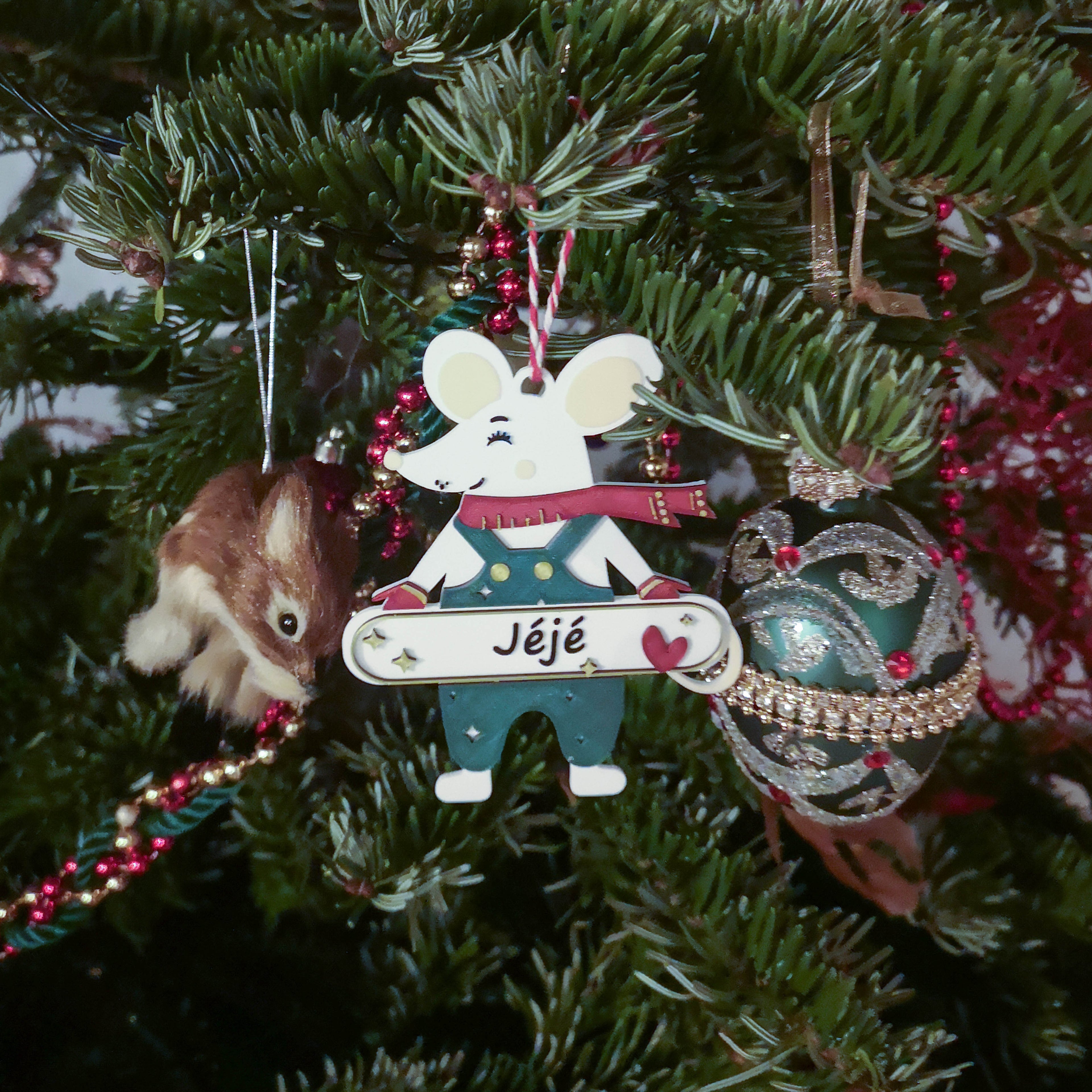 Décoration de Noël personnalisable en forme de souris, suspendue sur un sapin : souris avec écharpe rouge et salopette, tenant une pancarte avec prénom (ex. “Jéjé”) et un cœur, parmi guirlandes et boules de Noël.