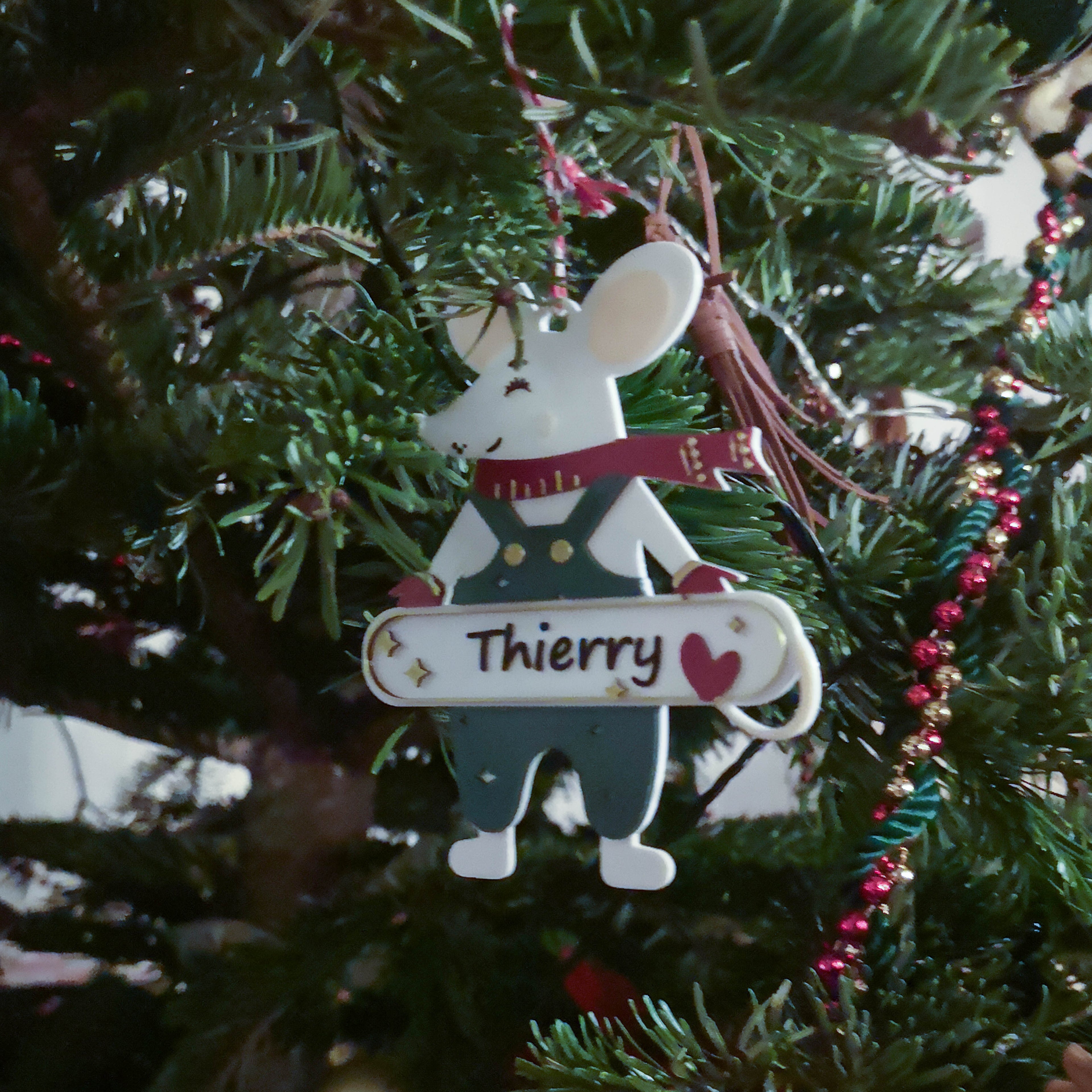 Décoration de Noël personnalisable en forme de souris, suspendue sur un sapin : souris avec écharpe rouge et salopette, tenant une pancarte avec un prénom (ex. “Thierry”) et un cœur, au milieu des branches du sapin.
