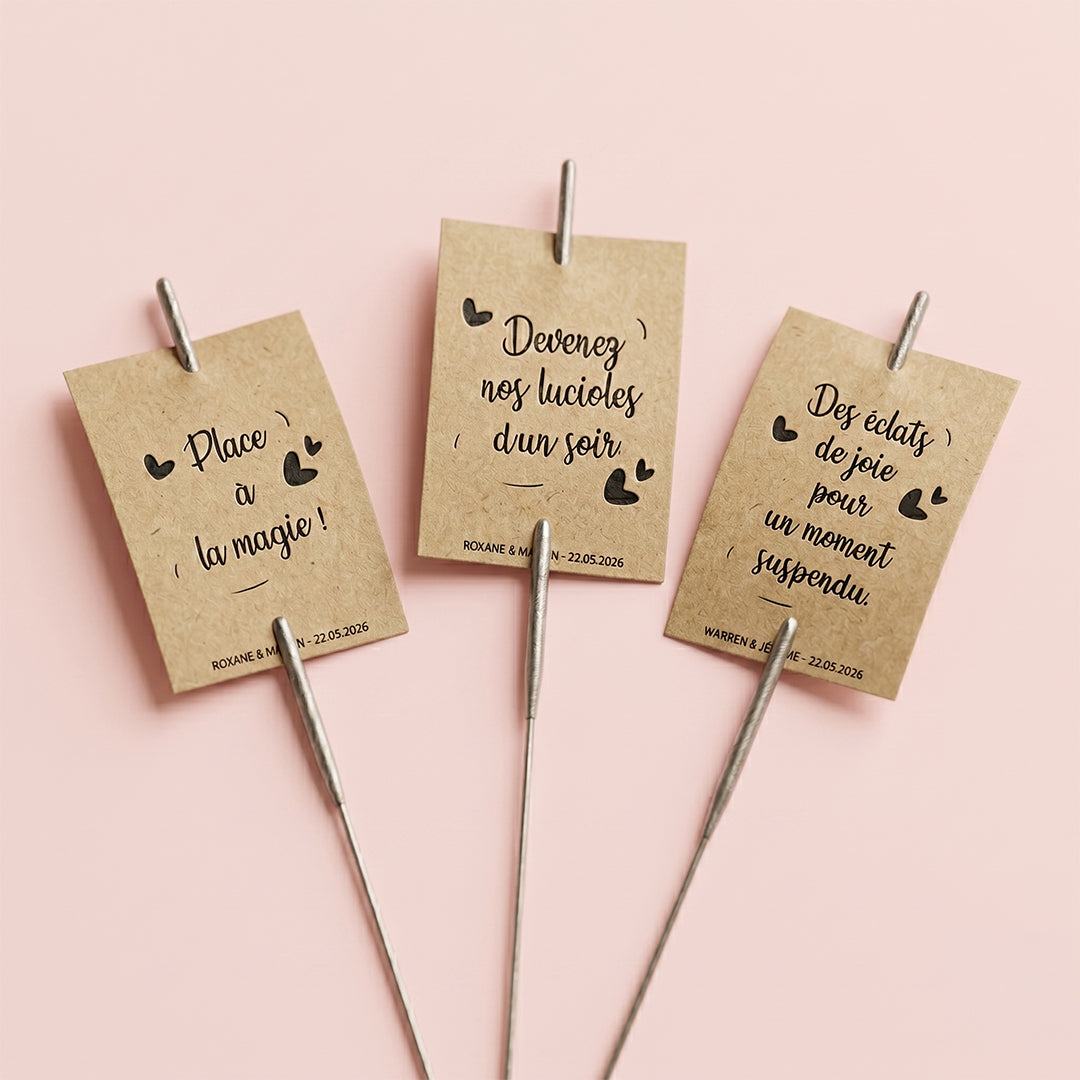 Trois étiquettes en papier kraft marron avec des messages personnalisés pour mariage (Place à la magie, Devenez nos lucioles d'un soir), fixées sur des cierges magiques métalliques sur fond rose pastel.