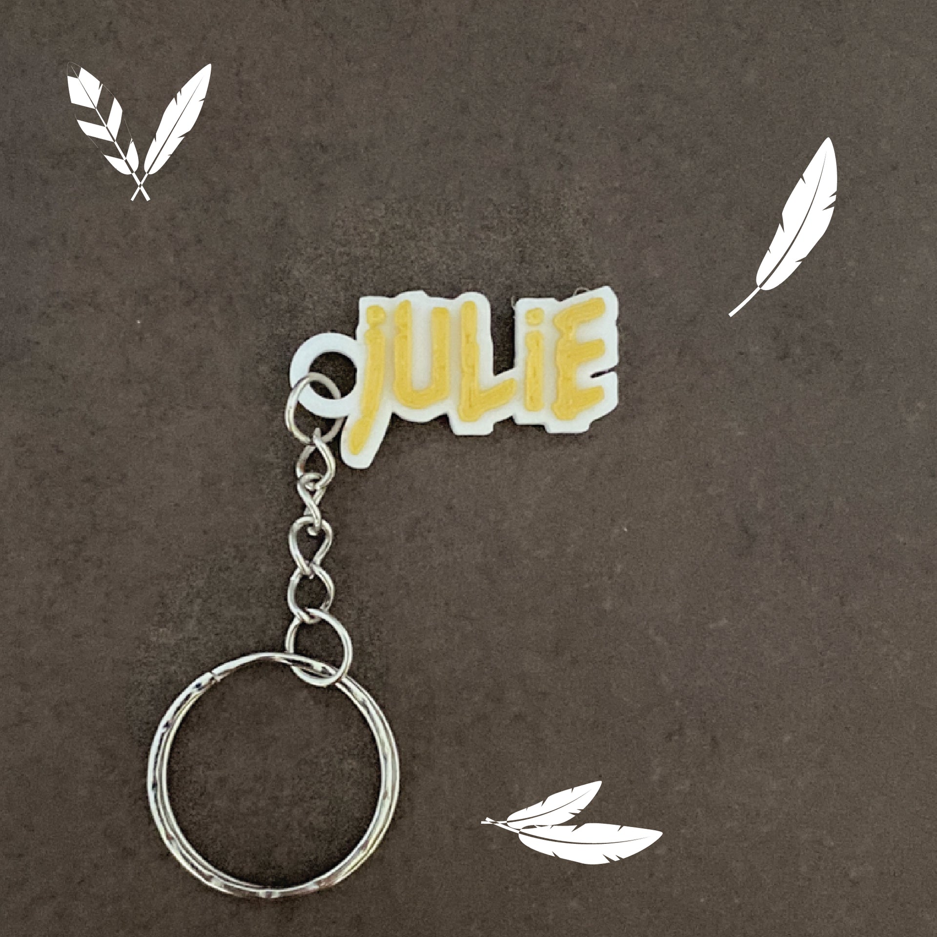 Porte-clés personnalisé au prénom Julie d'inspiration nature, style Pocahontas. Lettres dorées en relief sur support blanc texturé, décoré de plumes blanches stylisées sur fond gris foncé.