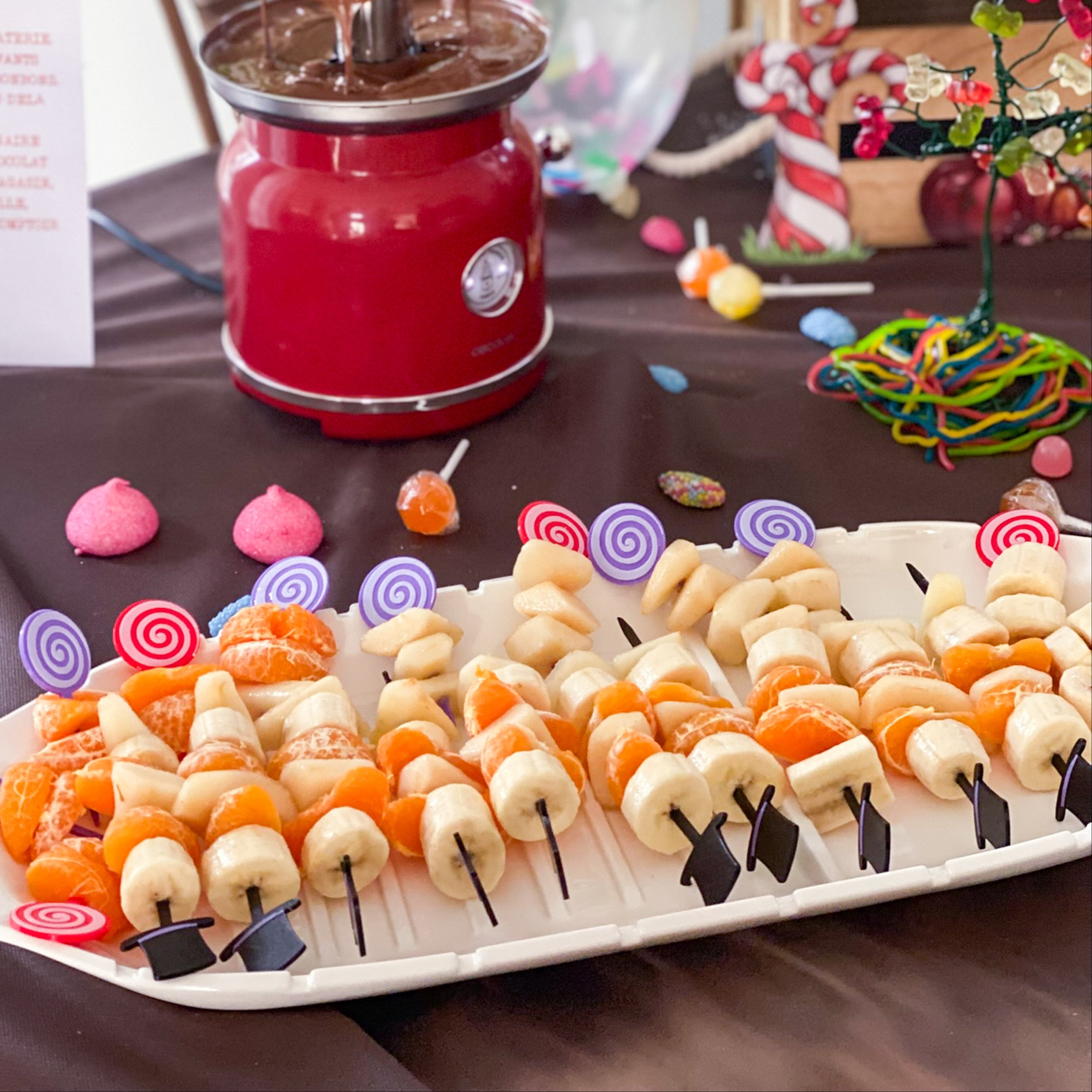 Vue d'ensemble d'un buffet festif avec un plateau de brochettes de fruits (banane et mandarine) décorées de piques "Chapeau Haut-de-forme" et "Sucettes Spirales". En arrière-plan, une fontaine à chocolat rouge et des décors colorés complètent cette table gourmande inspirée de l'univers de Charlie et la Chocolaterie.