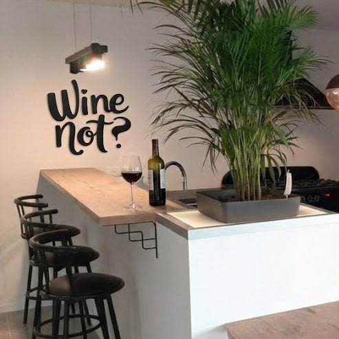 Décoration murale “Wine not?” en impression 3D (PLA) noire, installée dans un coin bar avec plan de travail en bois, tabourets, bouteille de vin et verres, sous une lampe murale et près d’une grande plante.