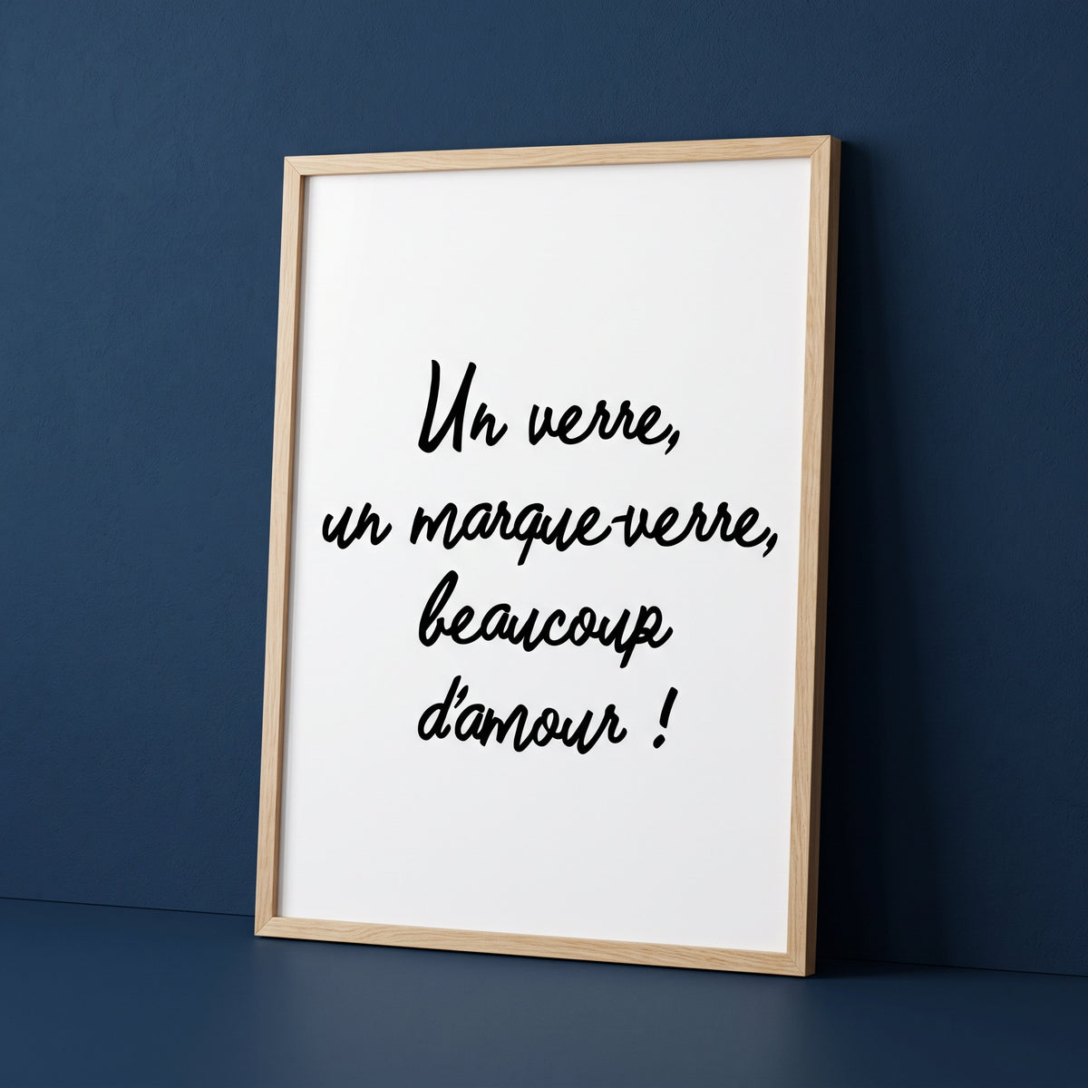 Affiche de décoration pour mariage, baptême ou fête, présentée dans un cadre en bois naturel, posée sur le sol contre un mur bleu foncé et texturé. L'affiche blanche, à orientation verticale, comporte le texte écrit dans une élégante police cursive noire : "Un verre, un marque-verre, beaucoup d'amour !". Ce panneau est conçu pour indiquer aux invités d'un événement (comme une réception de mariage ou un repas de baptême) de trouver et d'utiliser un marque-verre personnalisé pour leur verre.