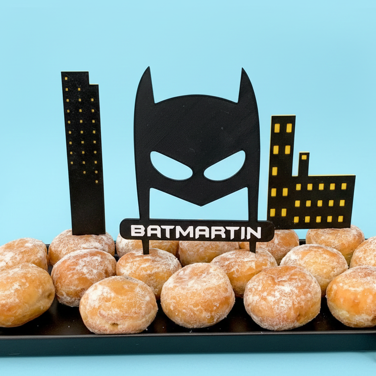 Cake topper personnalisé "BatMartin" en forme de masque de super-héros noir avec prénom, accompagné de décorations d’immeubles, posé sur un gâteau d’anniversaire pour enfant sur le thème des super-héros.