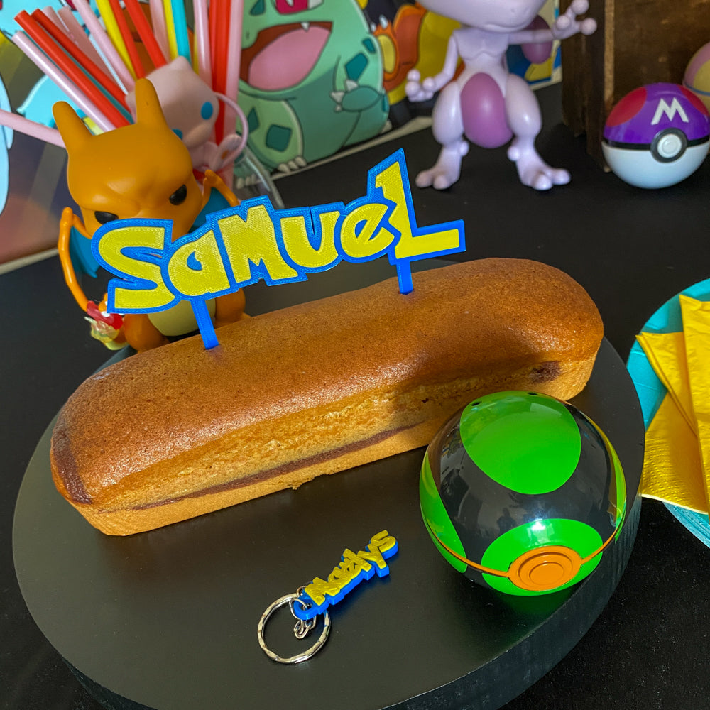 Pique à gâteau personnalisée 'SAMUEL' imprimée en 3D avec sa police type Pokémon et couleurs assorties bleu/jaune, présentée sur un cake. Sur le plateau noir devant le gâteau, deux porte-clés imprimés en 3D assortis sont posés : un avec le prénom 'SAMUEL' (même police et couleurs) et le porte-clés 'Maëlys' d'origine. L'ambiance est une fête d'anniversaire Pokémon avec des figurines Dracaufeu, Mewtwo, et une Poké Ball Friend Ball.