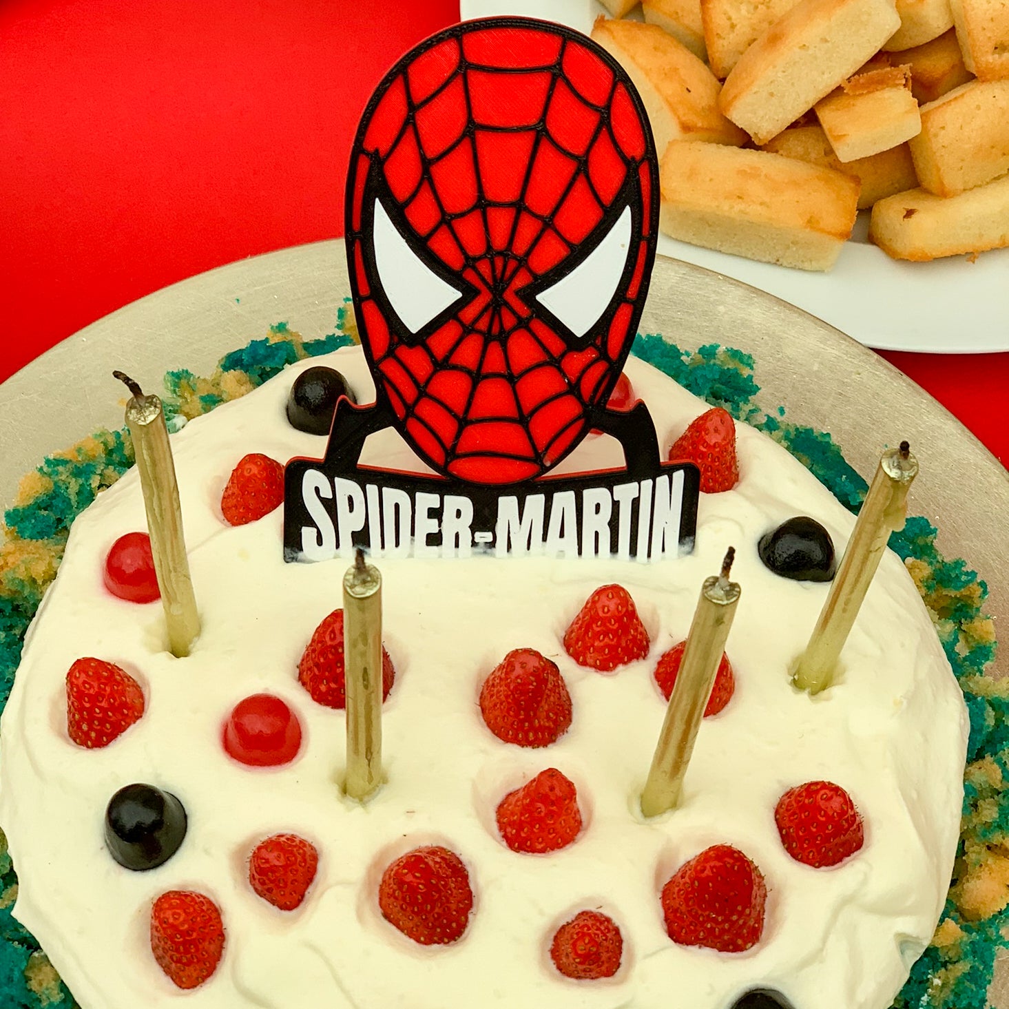 Cake d’anniversaire décoré avec un cake topper Spider-Man personnalisé, visage du super-héros rouge et noir et prénom Martin, posé sur un gâteau à la crème avec fruits rouges.
